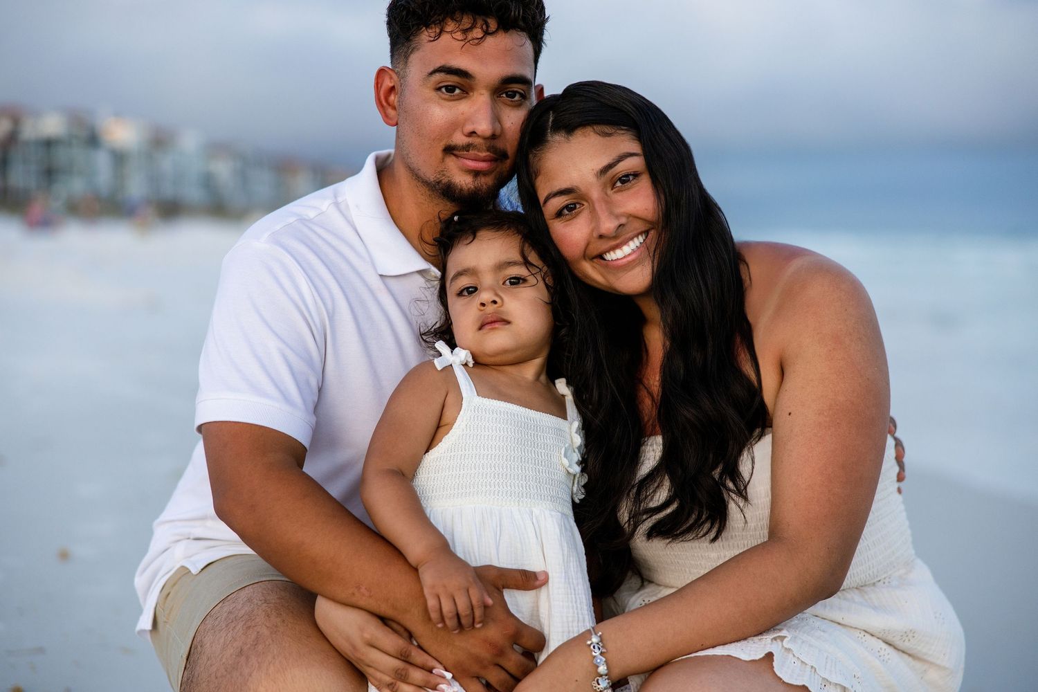 Young family of three poses on a sandy beach, wearing white outfits, smiling warmly on the beach in  pensacola, florida