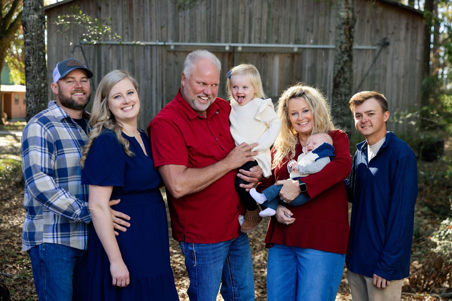 A joyful family of six poses outdoors near a rustic barn, wearing coordinated red and blue outfits, with a toddler held by grandparents.