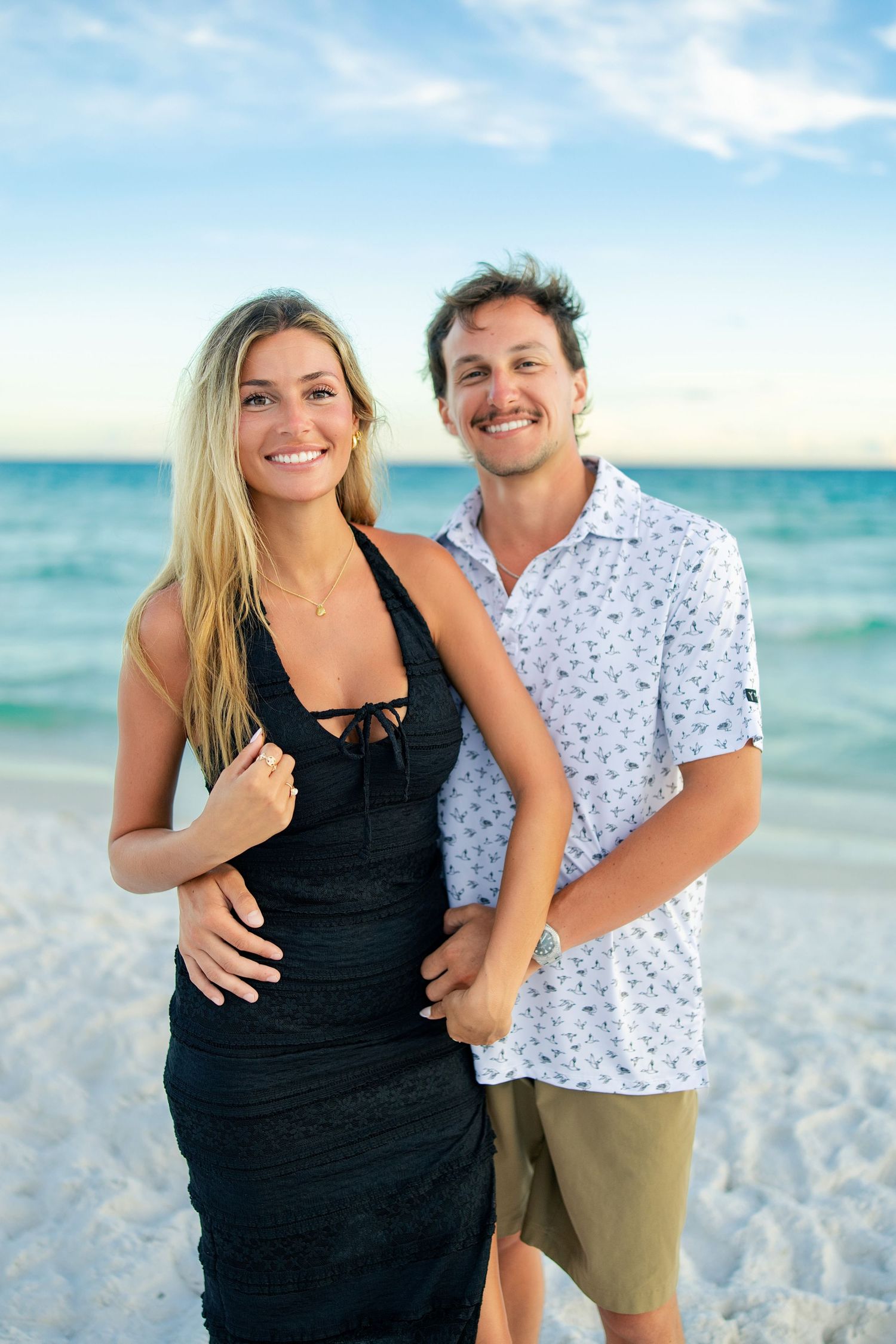 A smiling couple poses together on a white sand beach, woman in black dress, man in floral shirt and khaki shorts.