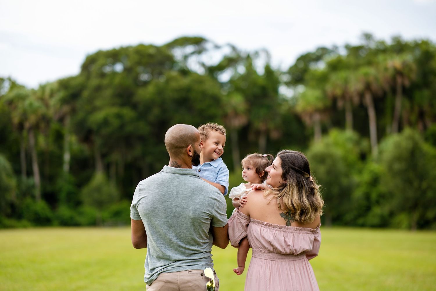 Family of four enjoying outdoor time together, parents holding young children in a lush green park setting.