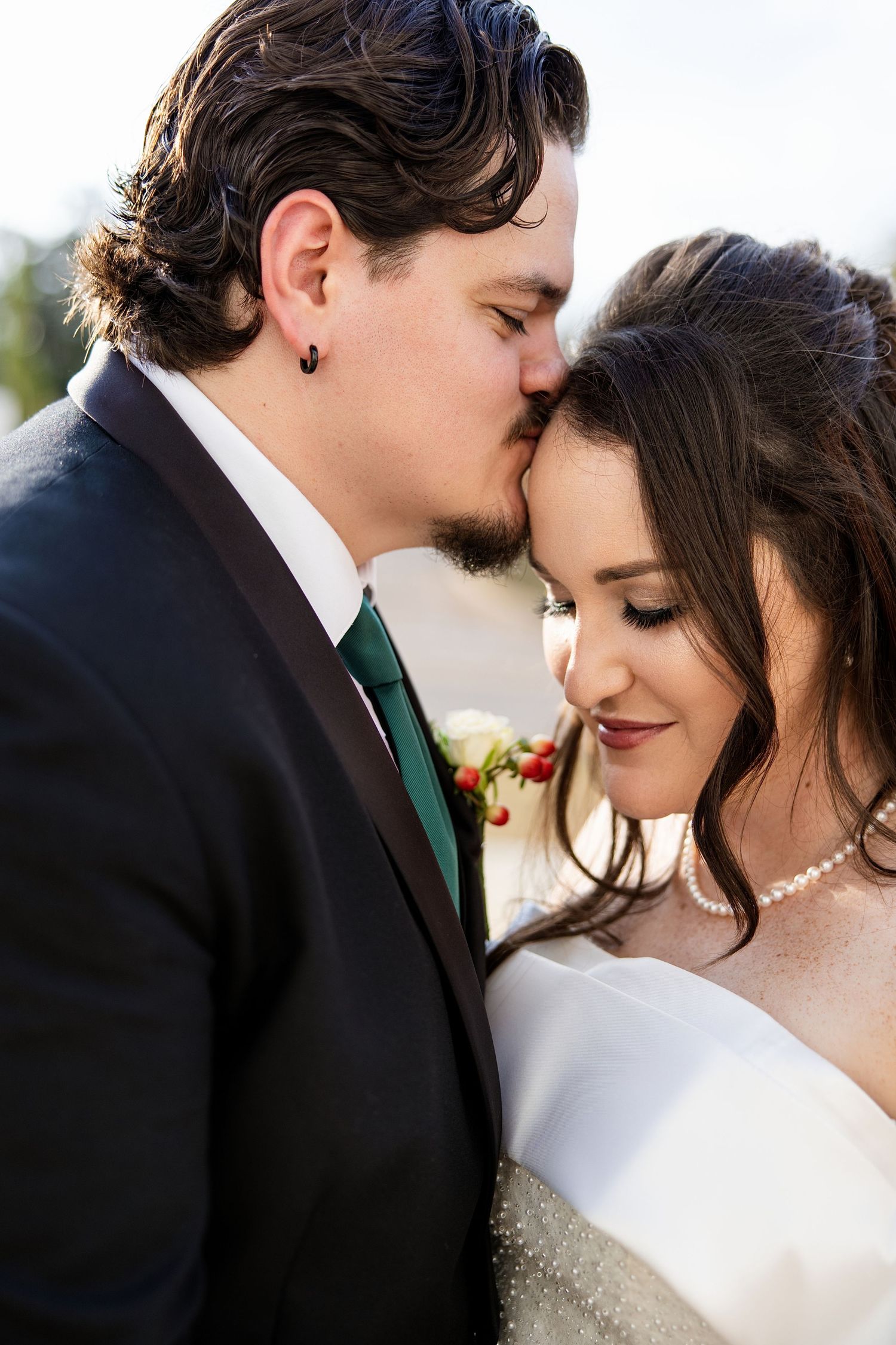 Groom in dark suit with green tie kissing bride on forehead, bride wearing pearl necklace and white gown.