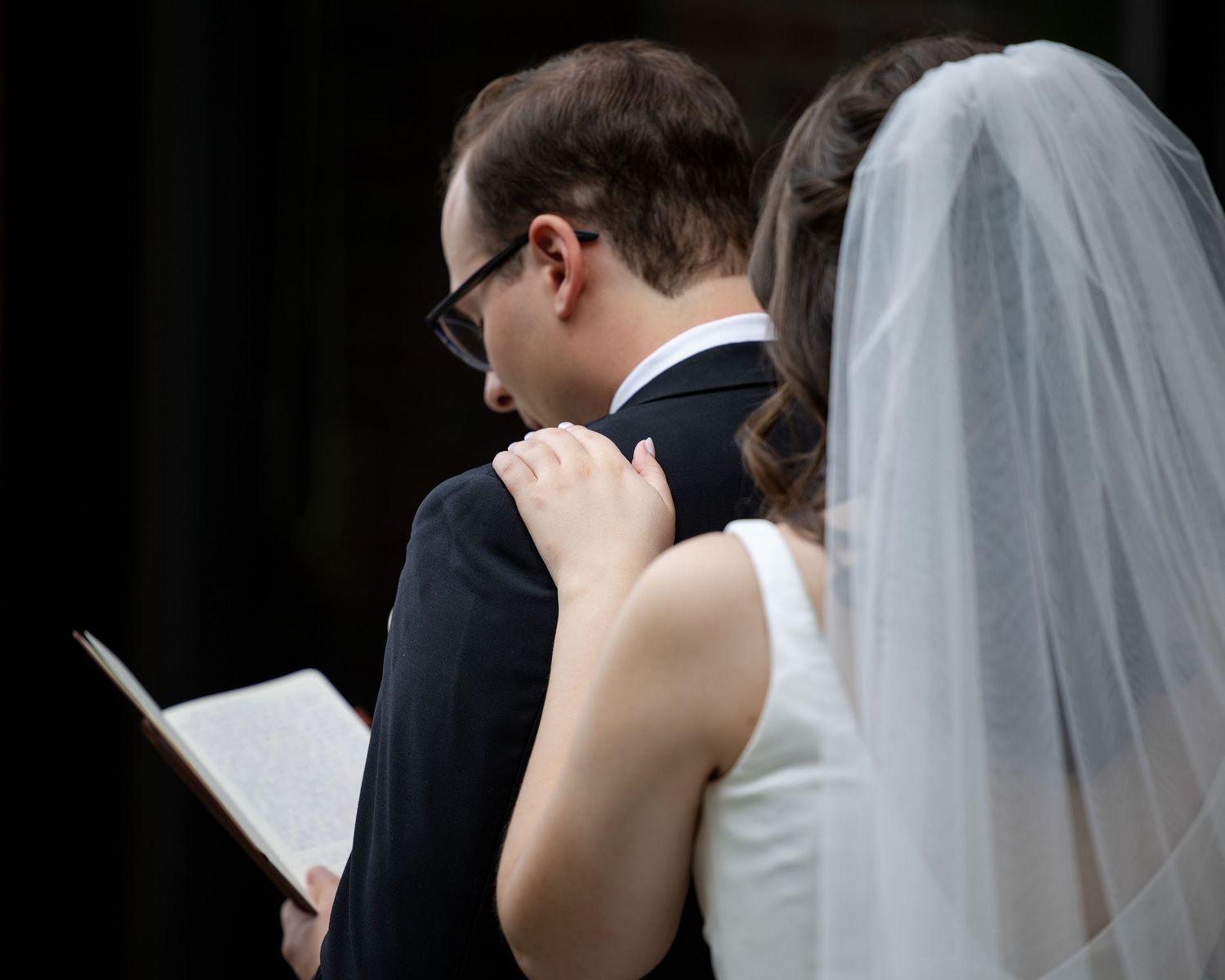 A groom in a black suit reads vows from a book as a bride in a white veil stands beside him at a wedding ceremony.