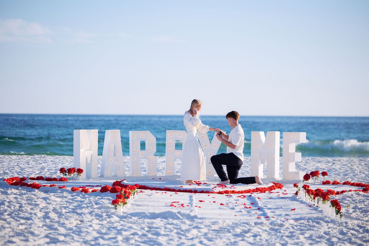 Beach proposal setup with large MARRY ME letters and rose petals on sand.