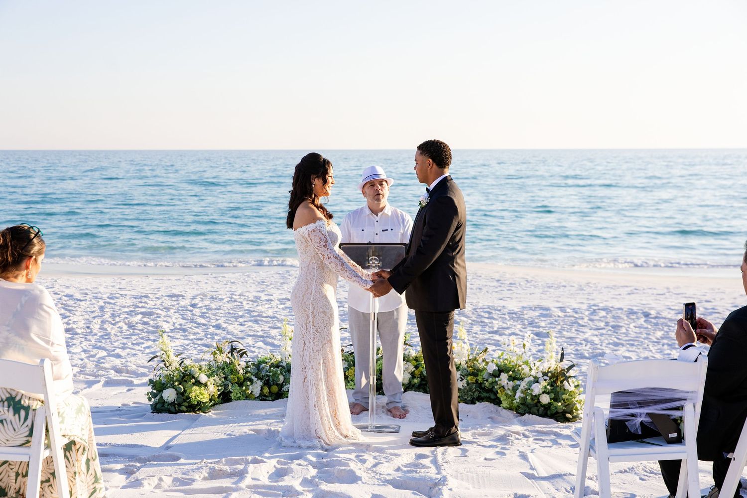 Couple exchanging vows at a romantic beach wedding ceremony with white floral arrangements and ocean backdrop.