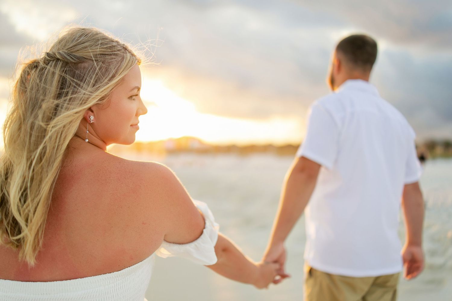 Couple holding hands on a beach at sunset, woman in white off-shoulder dress with blonde hair, man in white shirt.