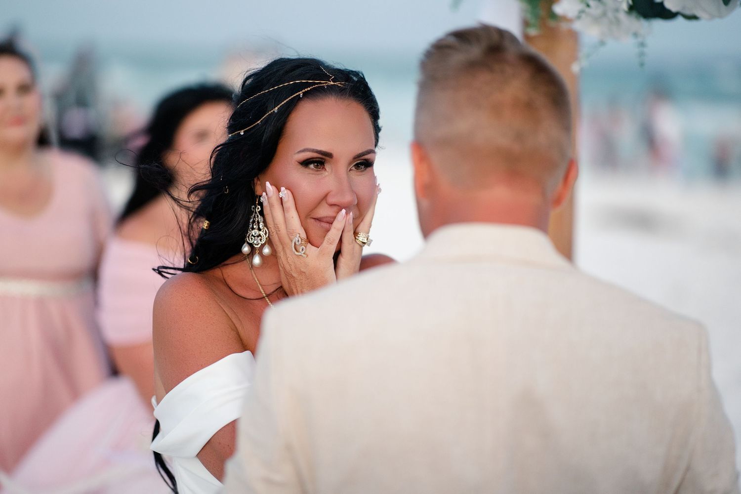 Emotional bride wiping tears during beach wedding ceremony with bridesmaids in pink dresses in background.