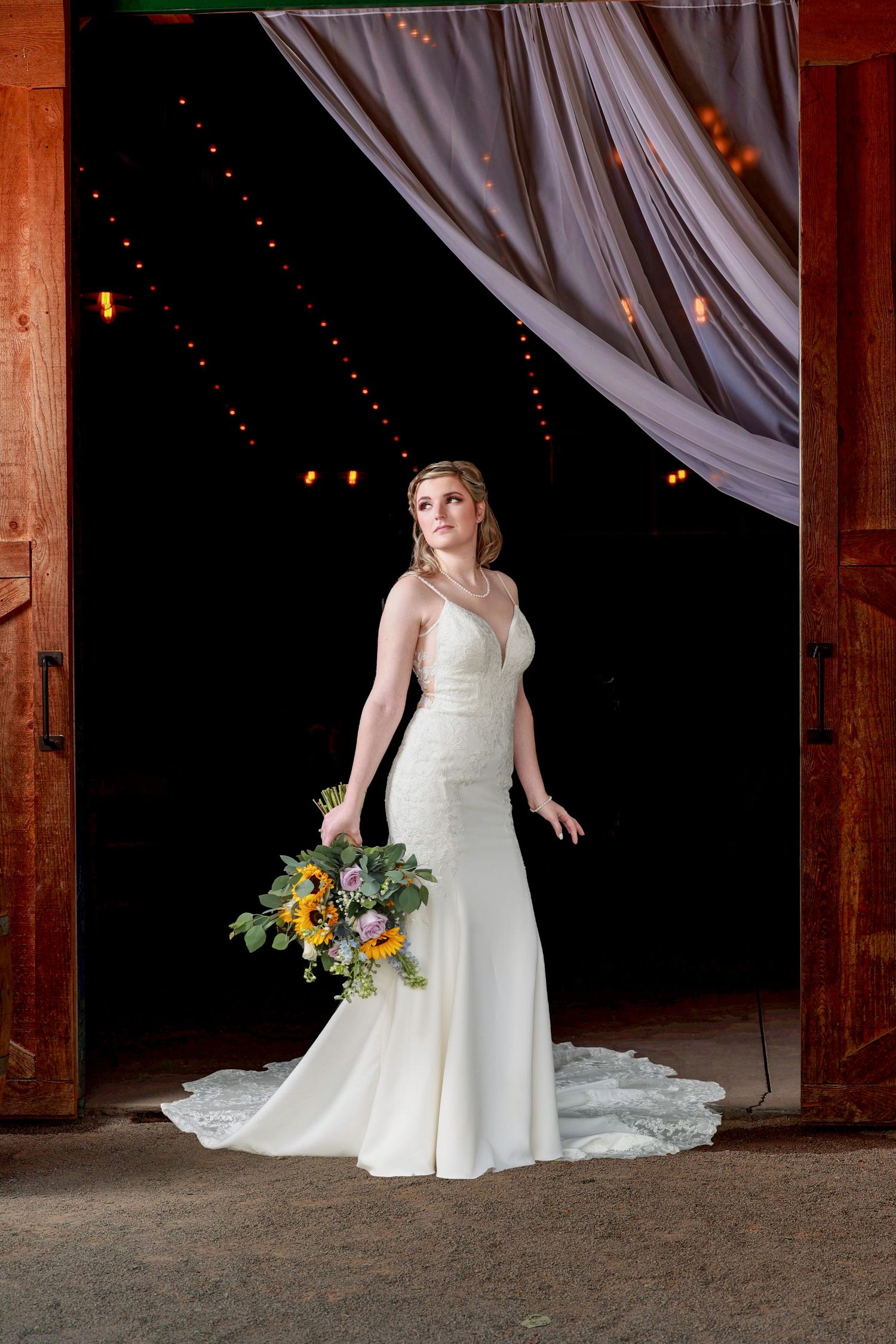 Bride in white mermaid gown holding sunflower bouquet stands in rustic barn doorway with string lights in pensacola, florida