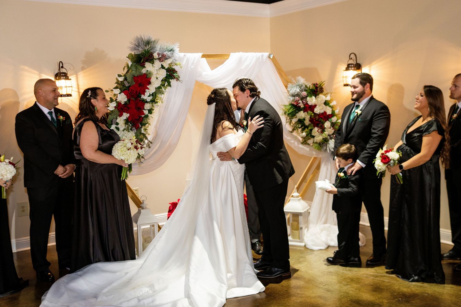 Bride and groom share first kiss at wedding altar decorated with red and white floral arrangements and draped white fabric.