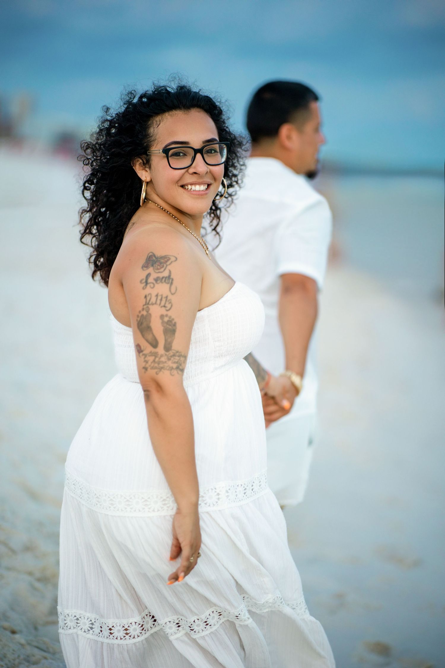 Smiling woman in white strapless dress with tattoos holds hands with man on sandy beach.