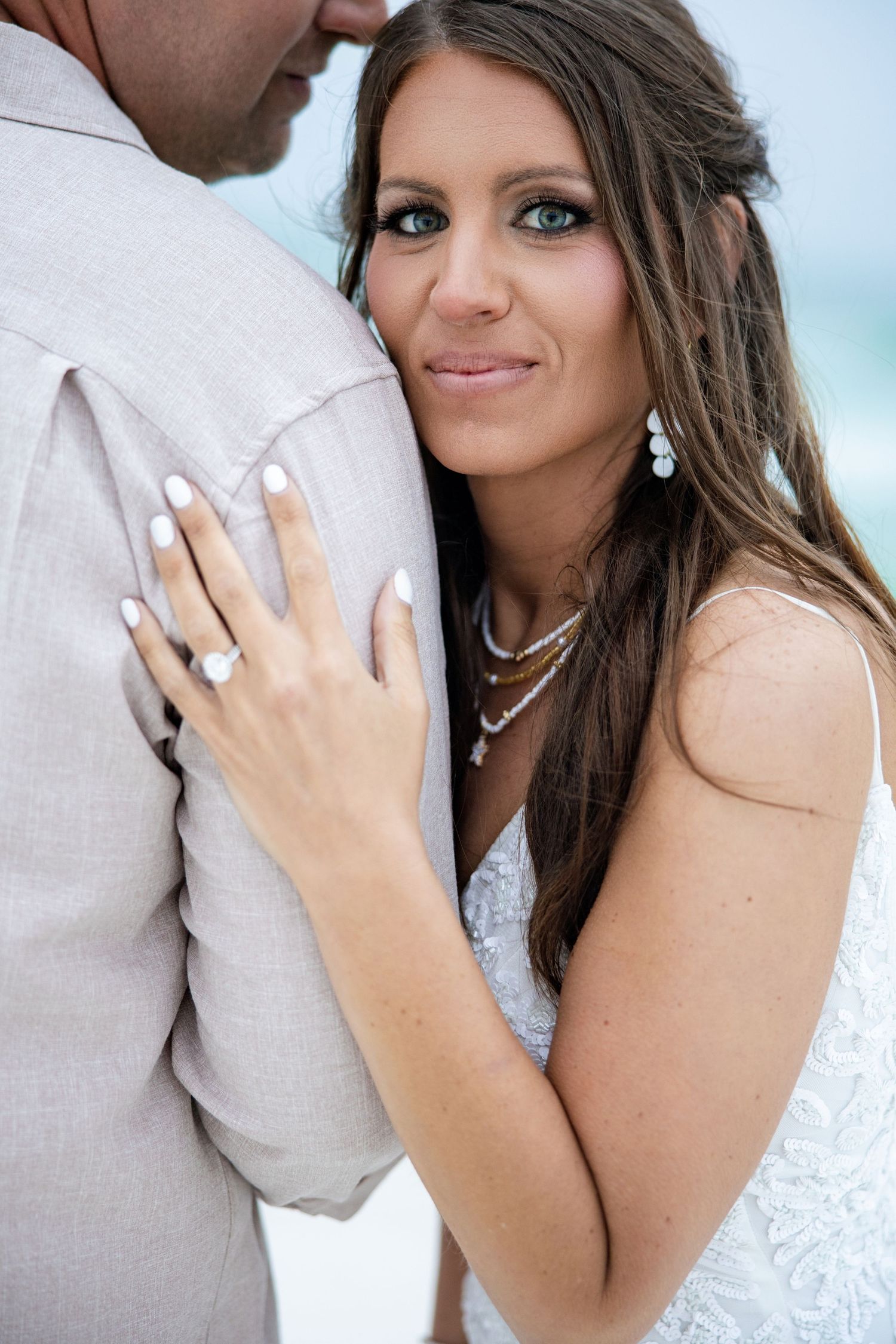 Bride in white lace dress with diamond ring and pearl jewelry leans against groom in tan suit at beach wedding.