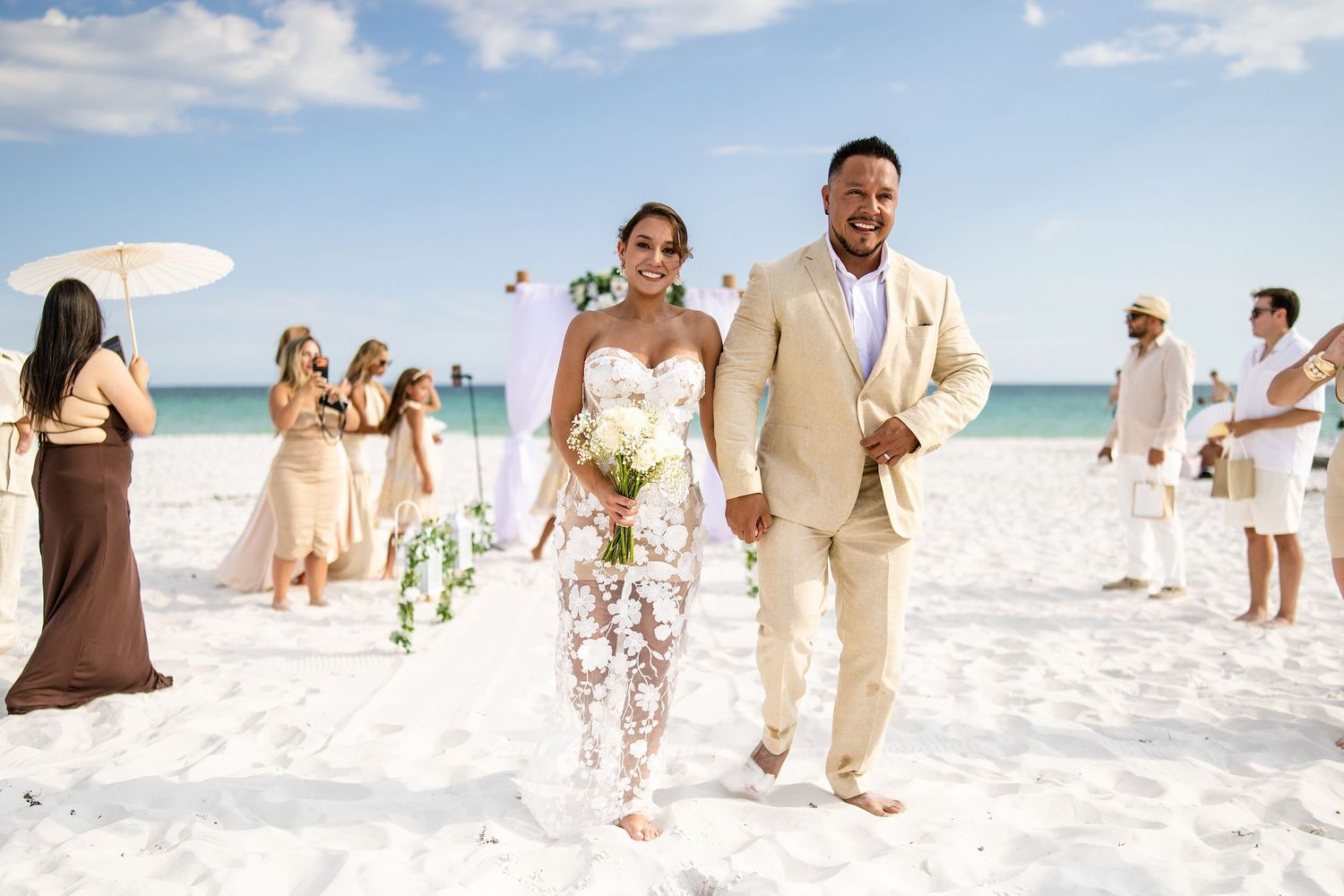 Couple walks hand-in-hand on white sand beach after wedding ceremony, bride in floral gown, groom in tan suit.