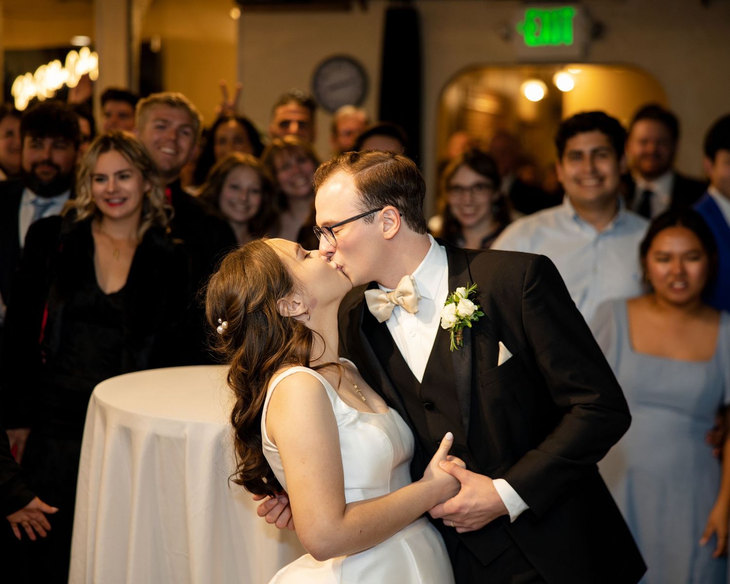 Bride and groom share first kiss at wedding reception surrounded by cheering guests in elegant venue.