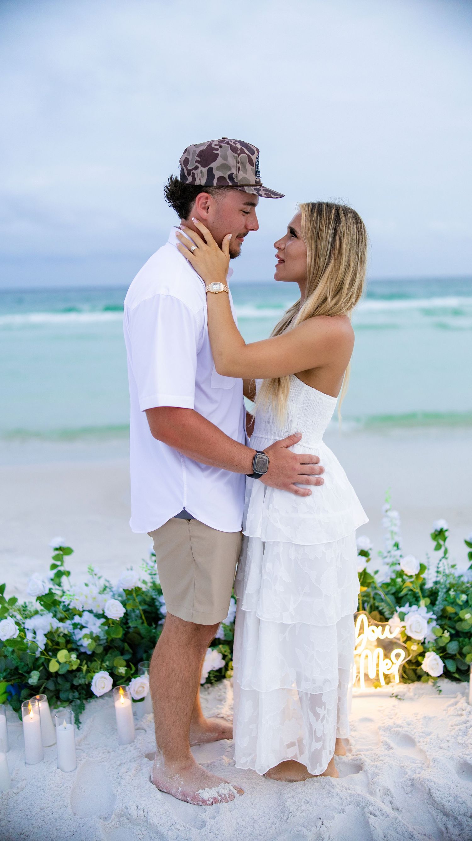 Couple in white outfits sharing a romantic beach proposal moment with floral decor and candles at sunset.