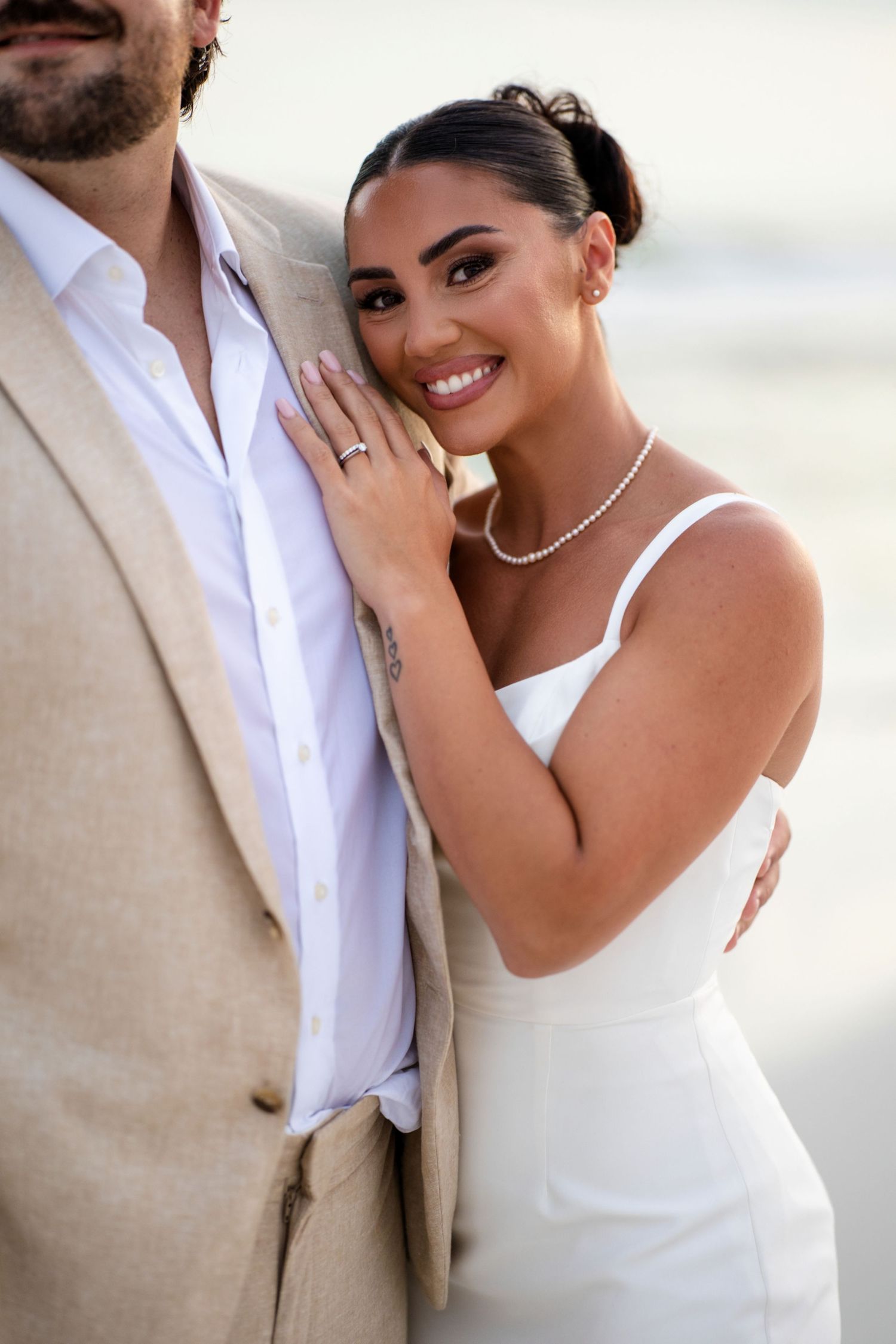 Smiling bride in white dress and pearl necklace leans against groom in tan suit during outdoor beach wedding photo.