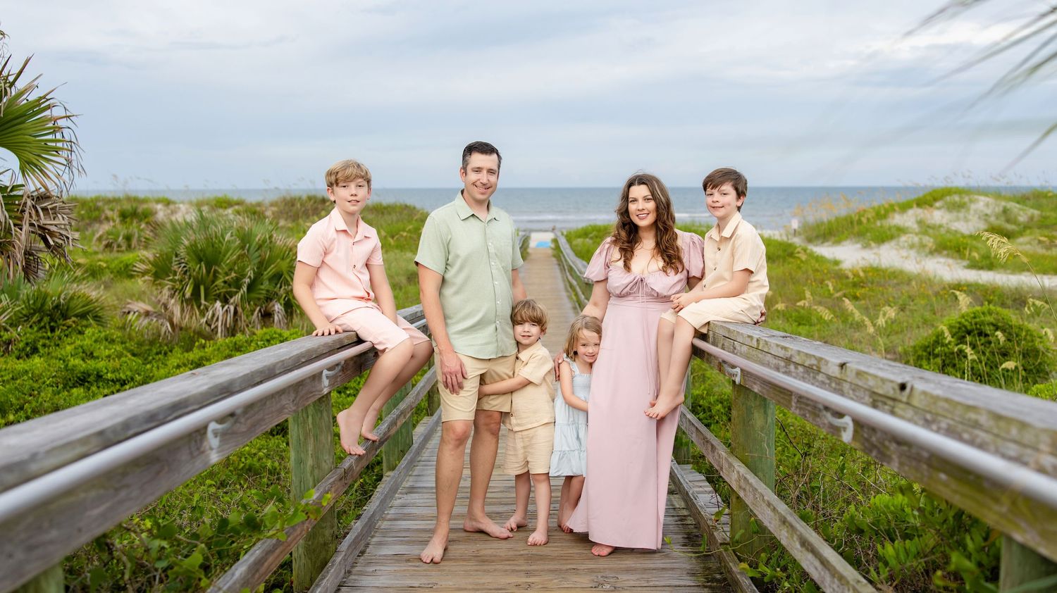 Family of five posing on a beachside boardwalk, dressed in coordinating pastel outfits with ocean views behind them.