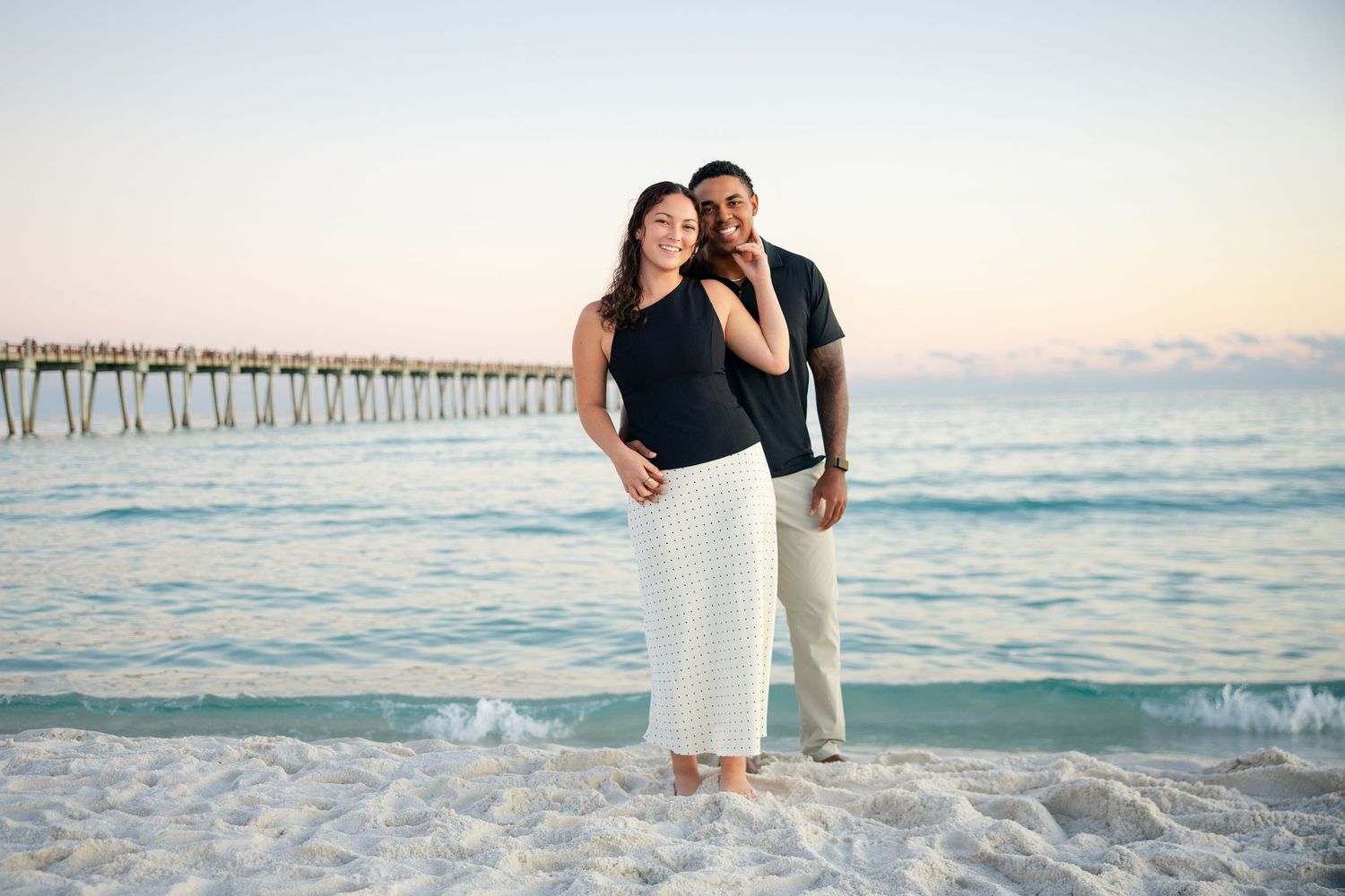 Couple standing together on a sandy beach near a pier at sunset, wearing casual black and white outfits.