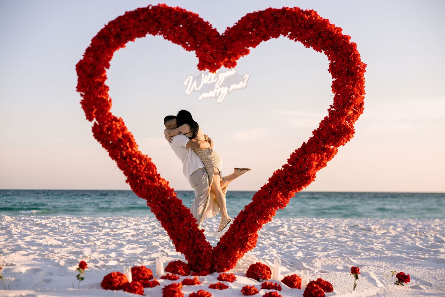 Couple kissing inside a large red rose heart arch on a beach at sunset during a romantic proposal.