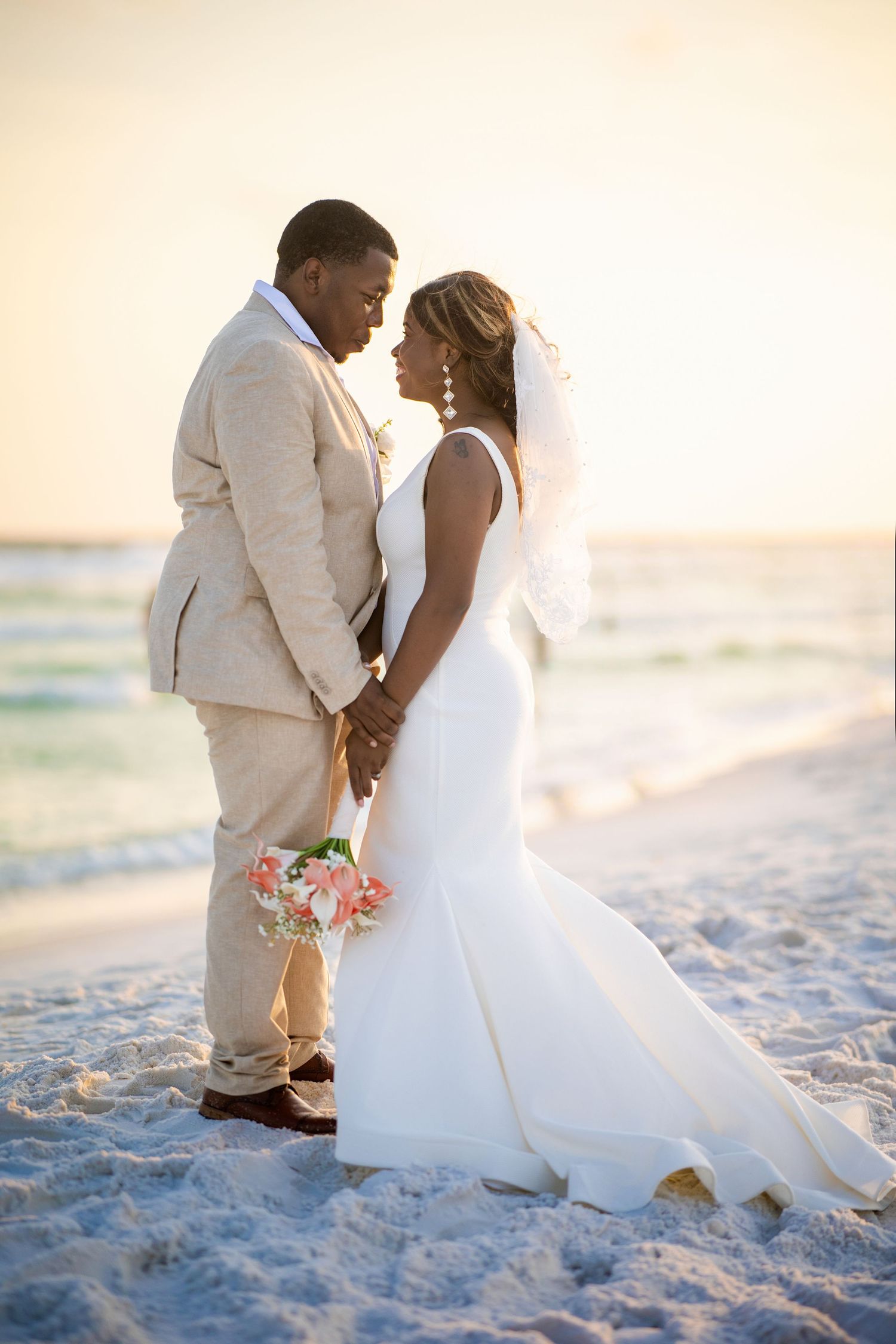 Newlywed couple holding hands on a sandy beach at sunset, bride in white gown with veil, groom in beige suit.