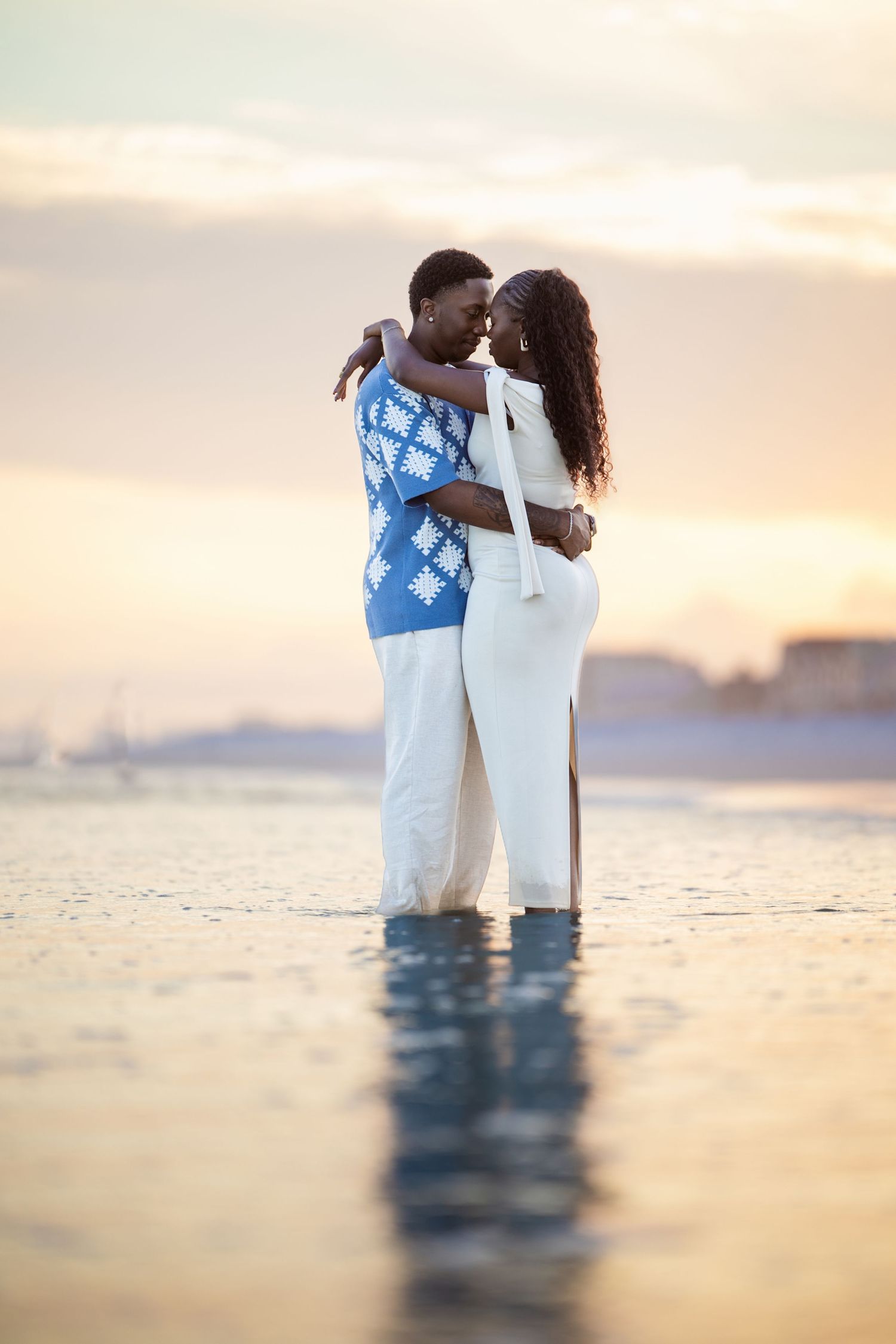 A romantic couple embraces in shallow ocean water at sunset, wearing white and blue traditional attire.