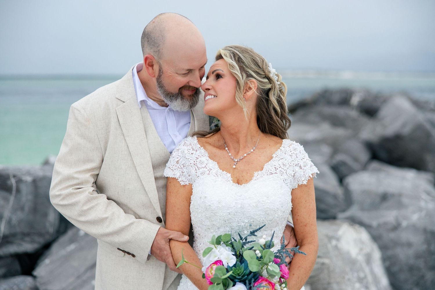 Bride and groom share a romantic moment by rocky shoreline, bride holding colorful bouquet with turquoise ocean backdrop.