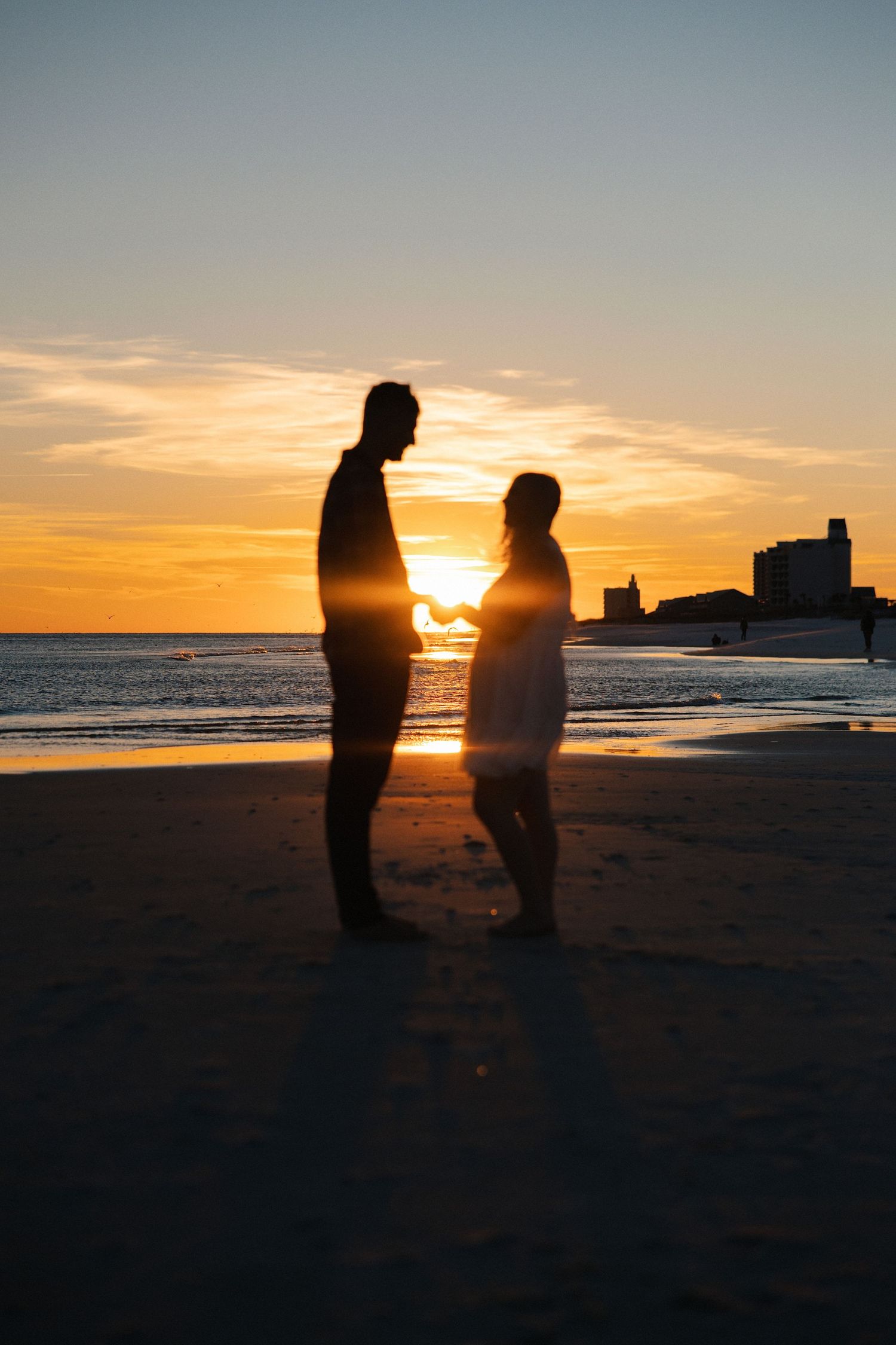 Silhouette of a couple holding hands on a beach at sunset with a golden sky and city skyline in the background.