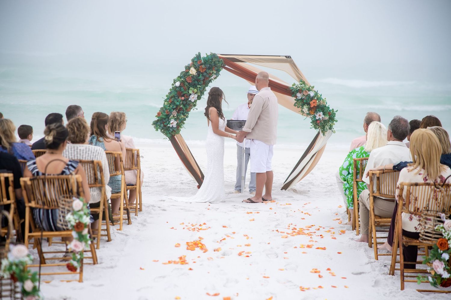 Romantic beach wedding ceremony with floral hexagonal arch, ocean backdrop, rose petal aisle, and seated guests on wooden chairs.