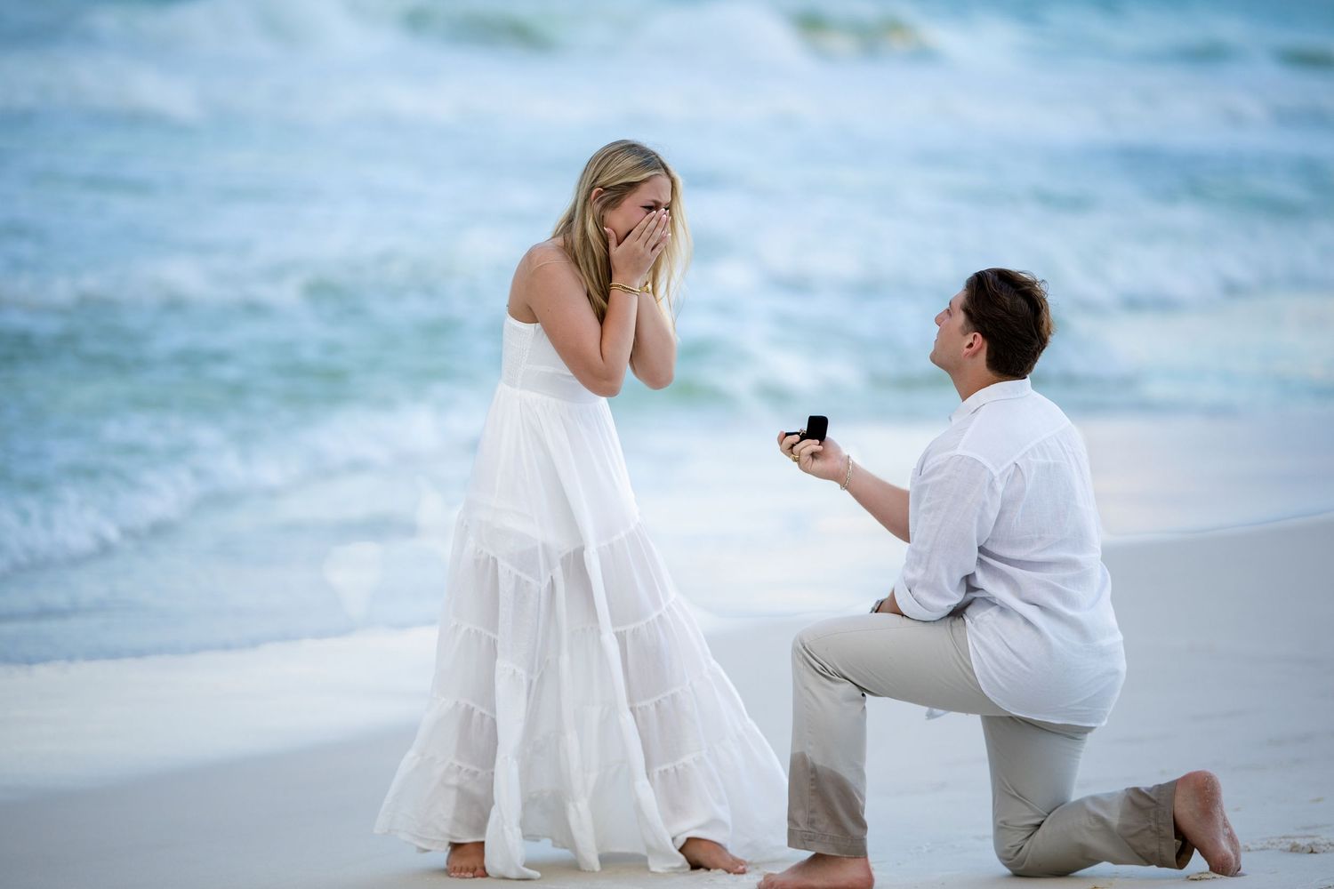 Emotional beach marriage proposal with man on one knee holding a ring box.