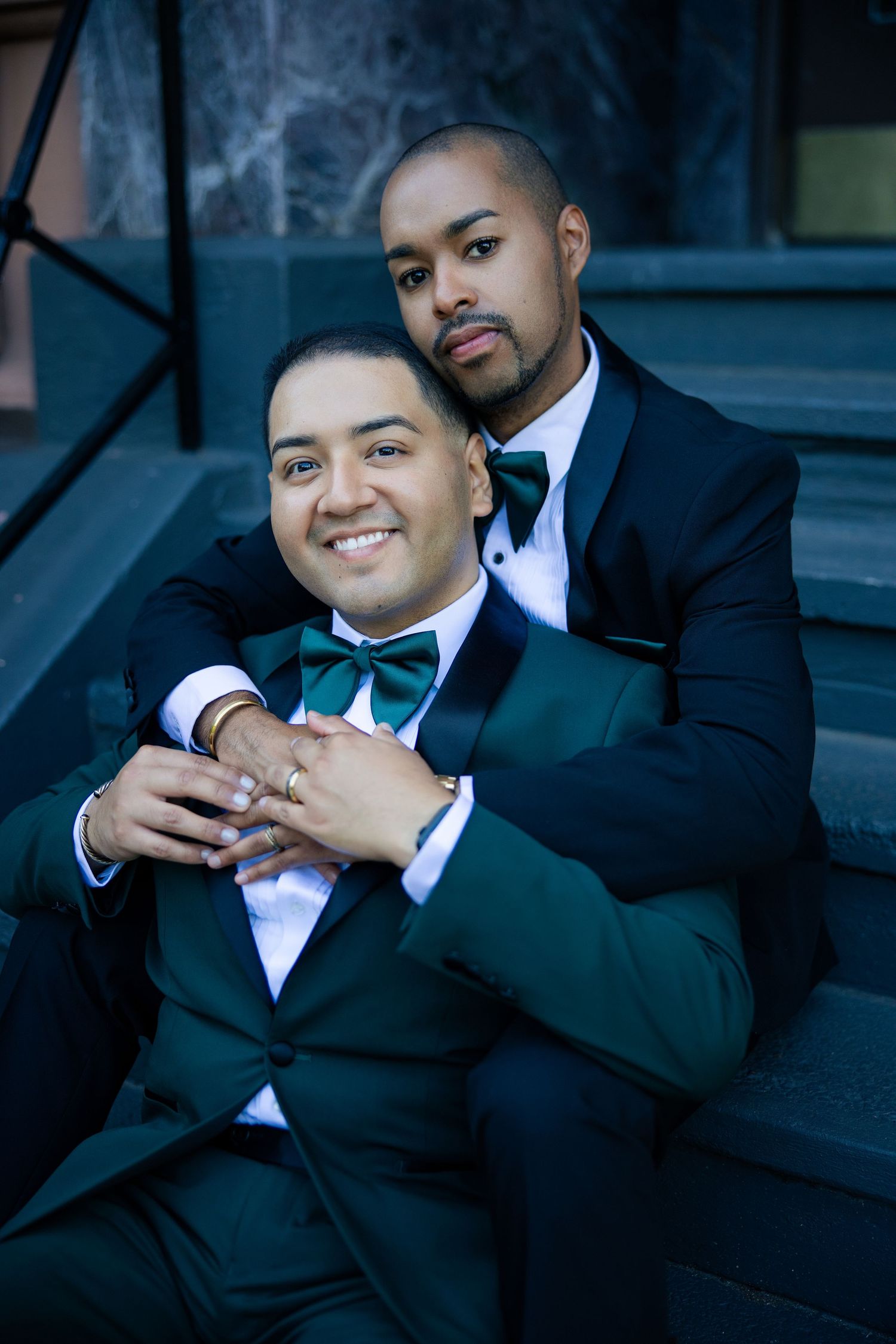 Two men in elegant green and black tuxedos pose together on steps, one embracing the other from behind.
