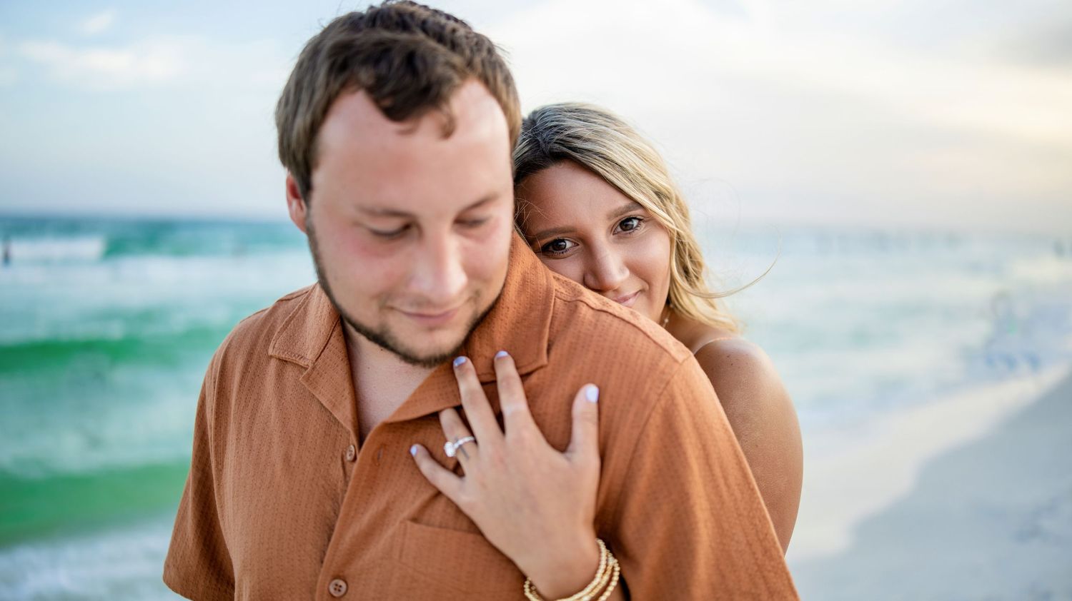 Couple embracing on a beach at sunset, woman showing engagement ring while hugging man in brown shirt.