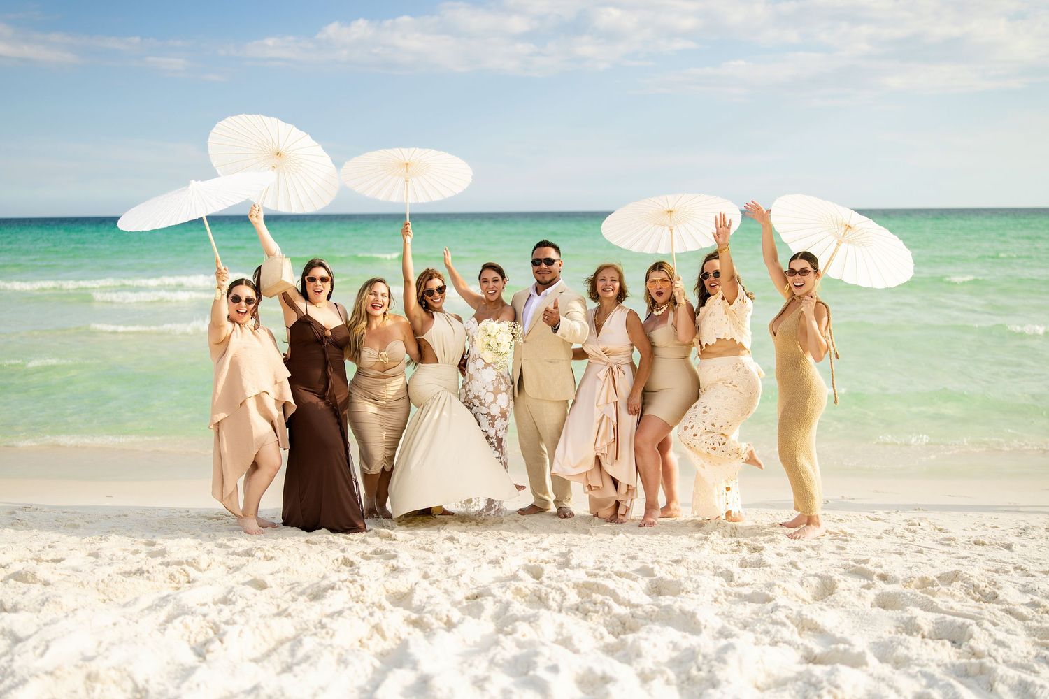 Wedding party celebrating on a sunny beach, bride and groom with bridesmaids holding white parasols, turquoise ocean backdrop.