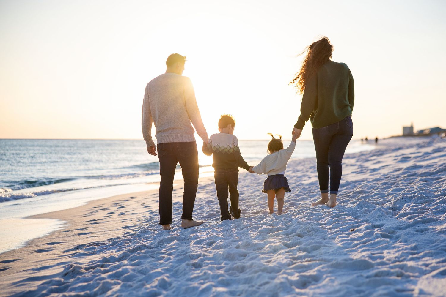 Family of four walking hand-in-hand on a sunny beach at sunset, with waves gently lapping the shore.