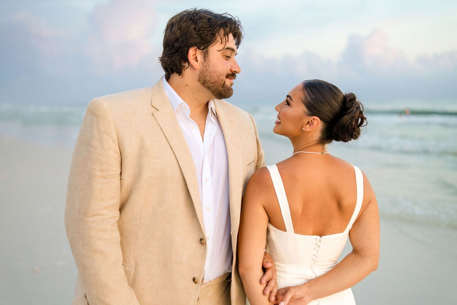 Couple in wedding attire sharing a romantic moment on a beach at sunset, groom in tan suit, bride in white dress.