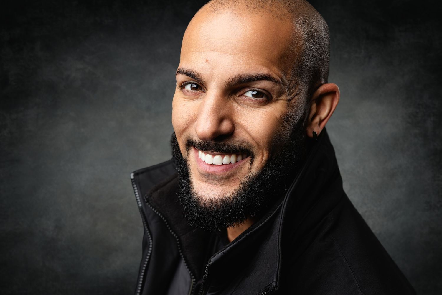 Smiling bald man with a beard wearing a black jacket, posing for a professional portrait against a gray background.