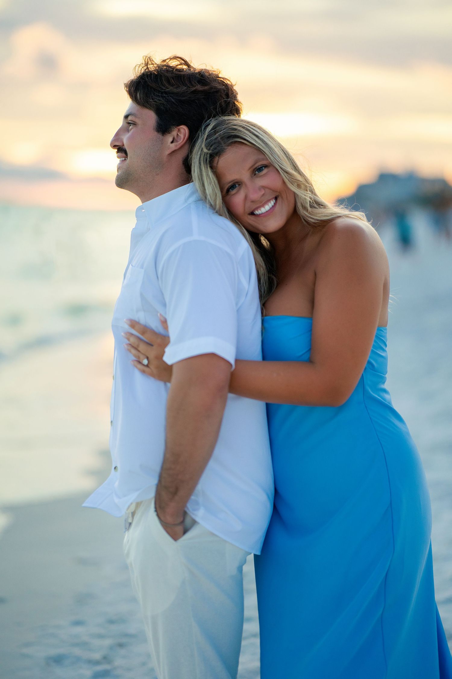 Couple embracing on a beach at sunset, woman in blue dress smiling, man in white shirt, ocean backdrop.