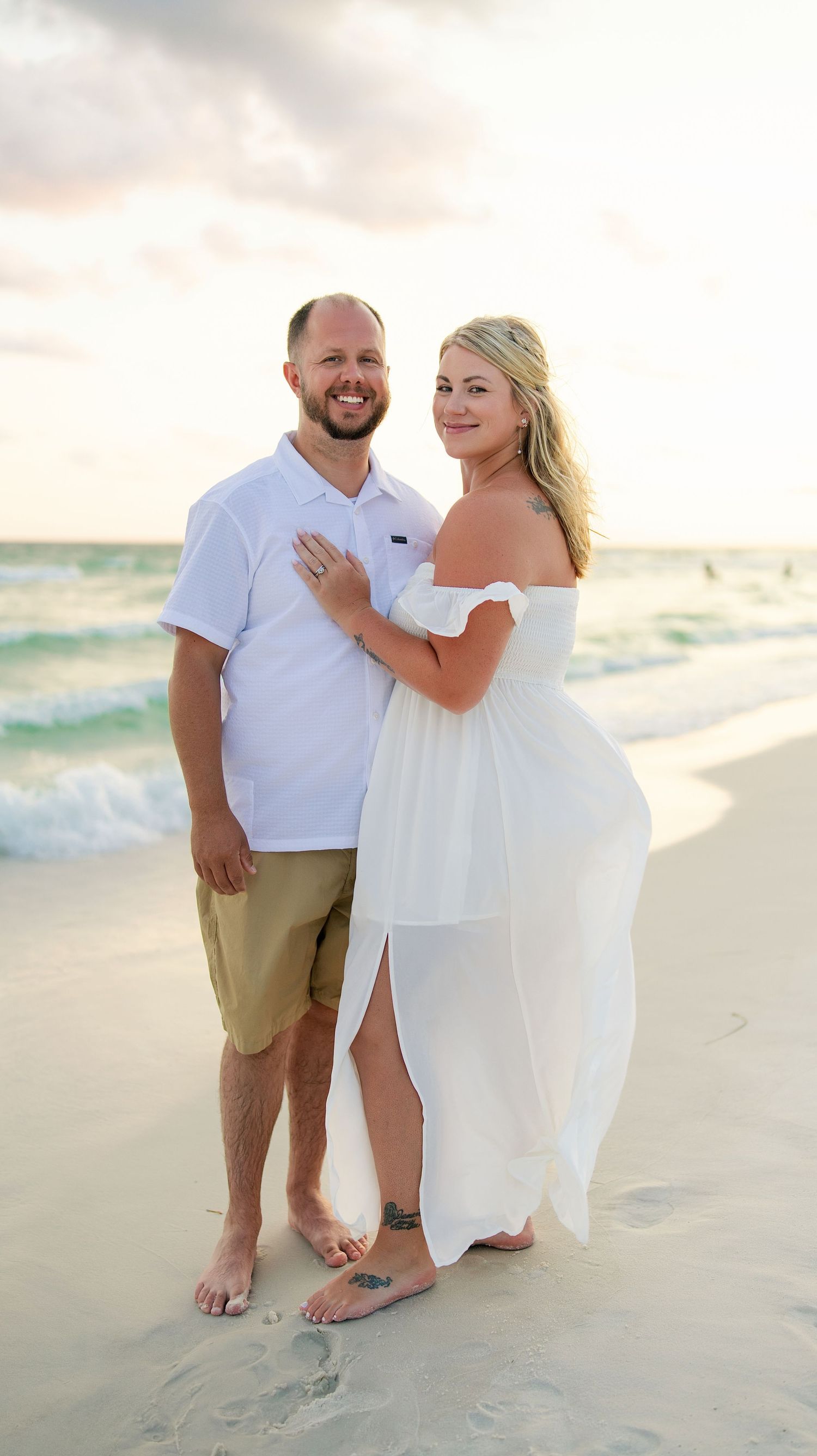 Couple in white outfits posing on a sandy beach at sunset with ocean waves in the background.