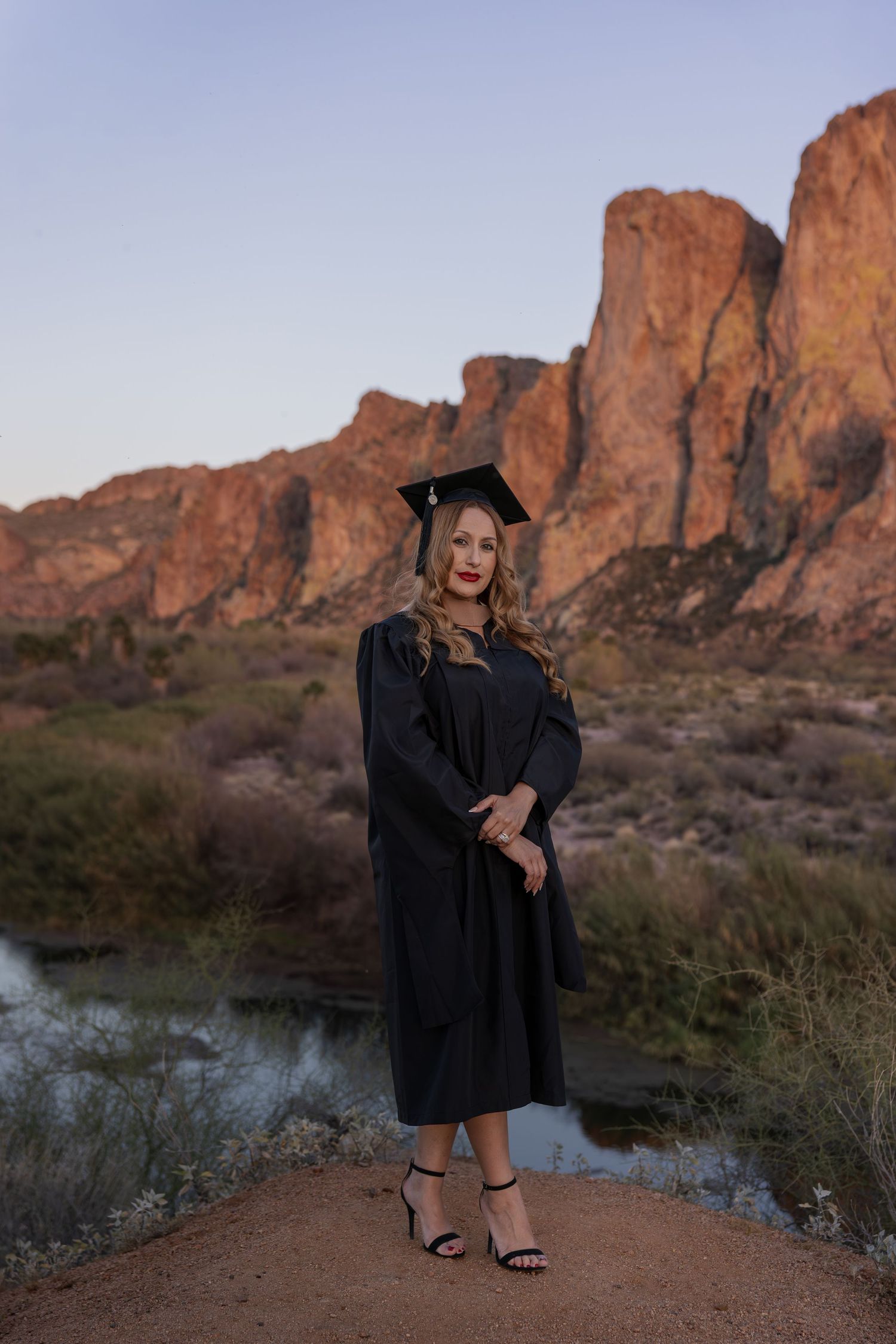Graduate in black cap and gown poses proudly on a desert path with stunning red rock formations at dusk.