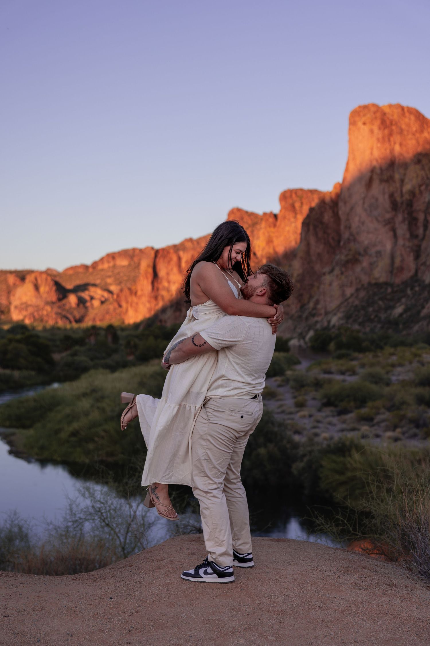 Couple embracing at sunset with glowing red rock mountain and river in desert landscape.