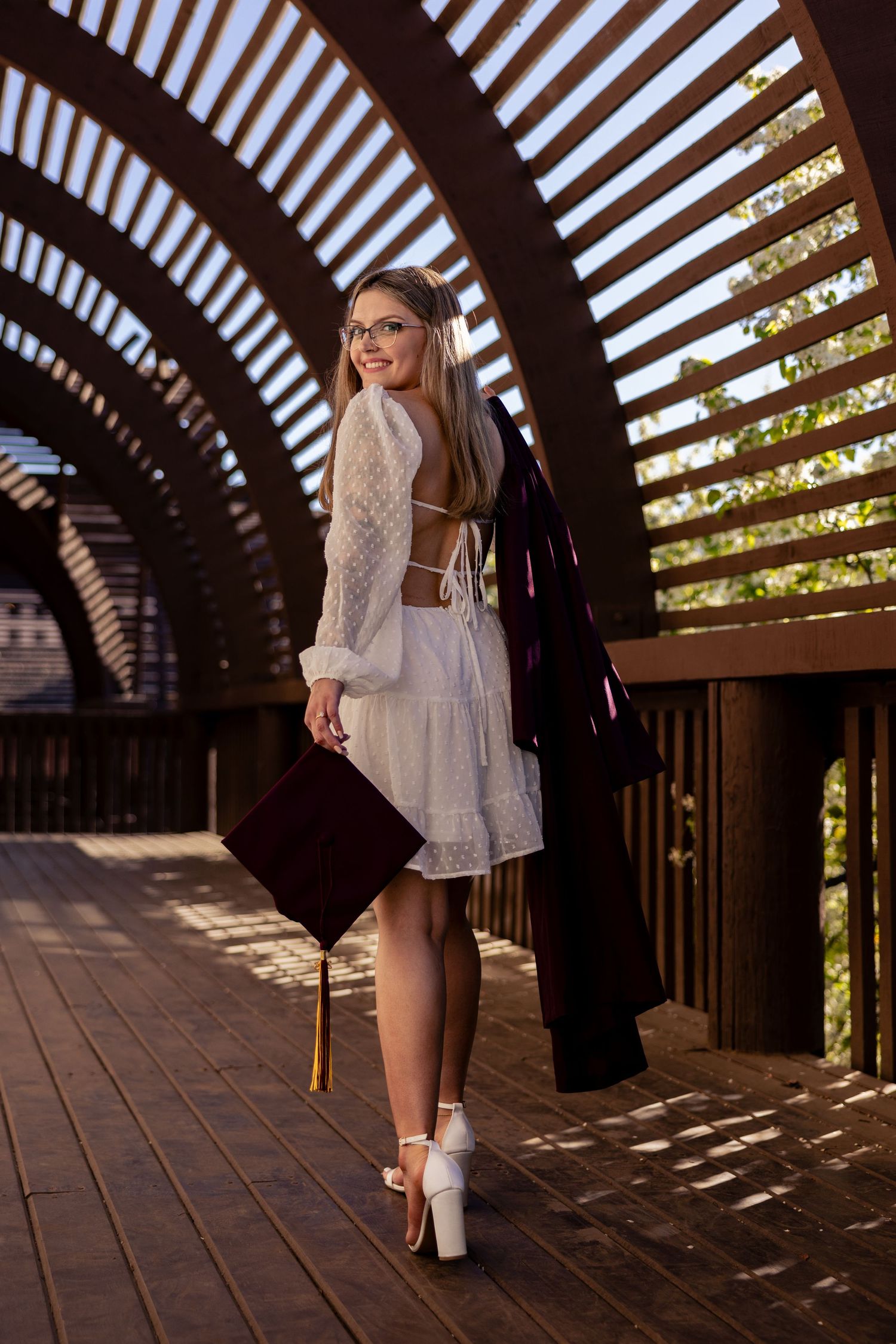 Smiling graduate in white dress carries cap and gown through decorative wooden arch tunnel.