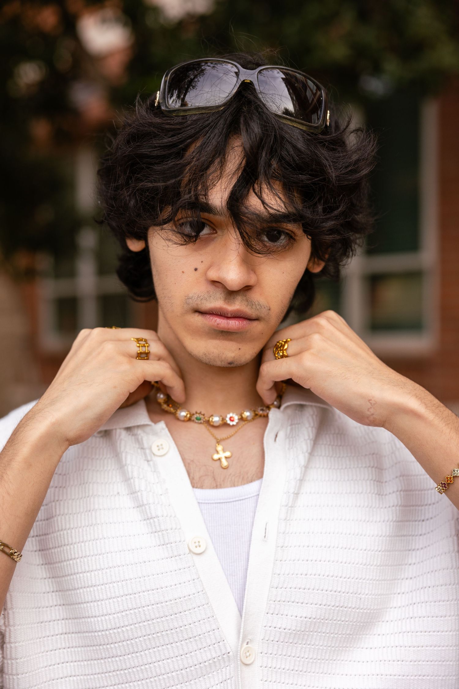 Young man in white shirt adjusting collar wearing gold jewelry and sunglasses outdoors.