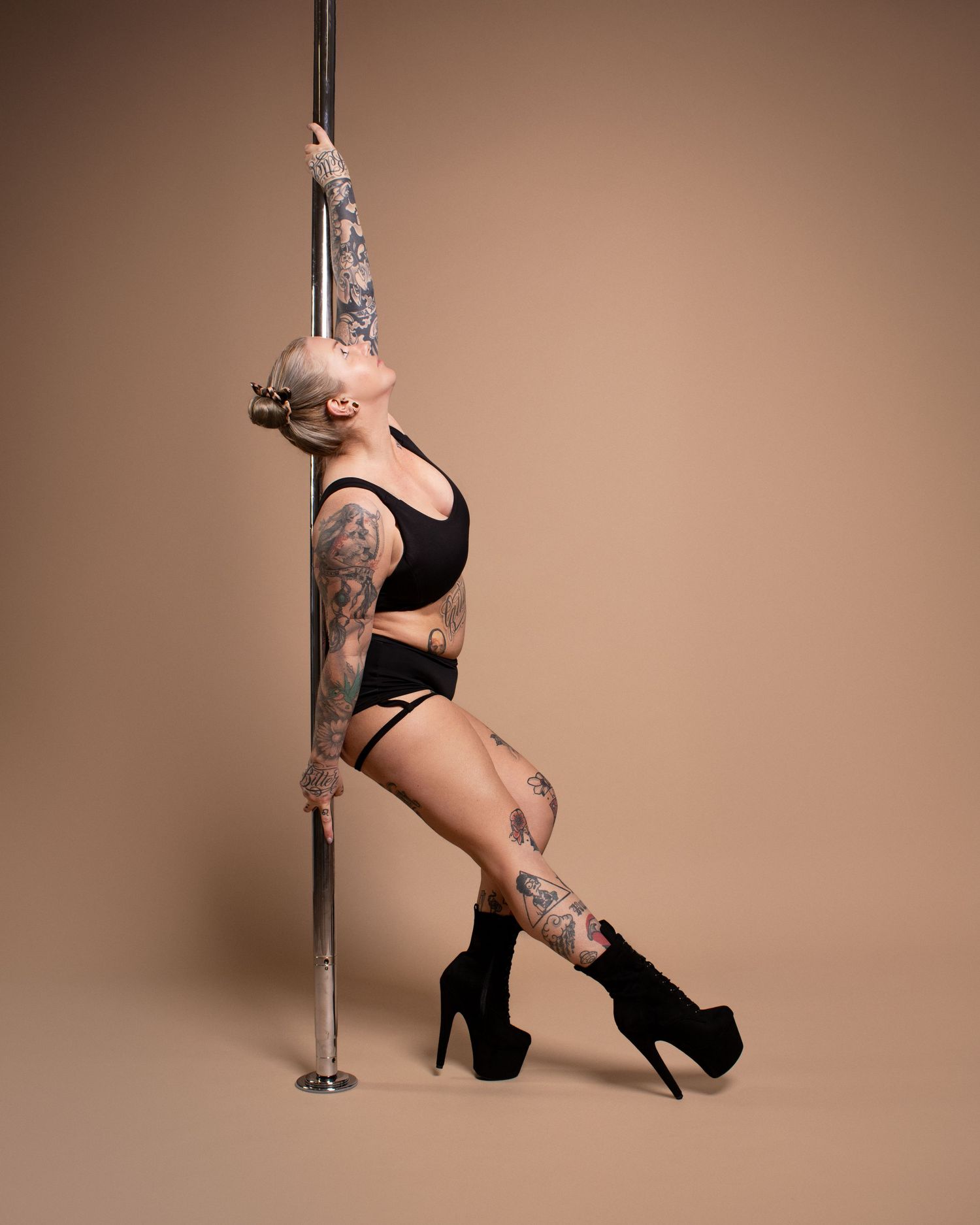 Tattooed woman in black lingerie and heels poses gracefully against a pole on a beige studio background.