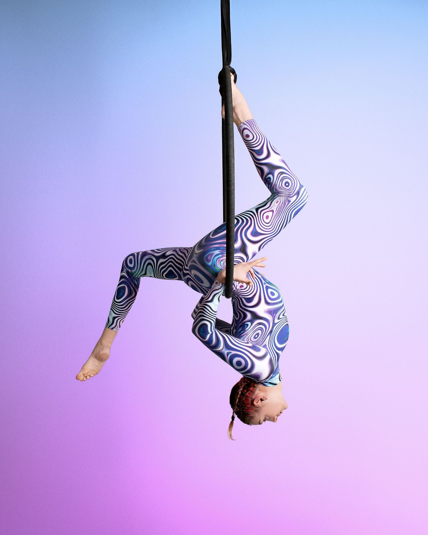 Aerial performer in a patterned bodysuit gracefully hangs upside down against a purple gradient background.