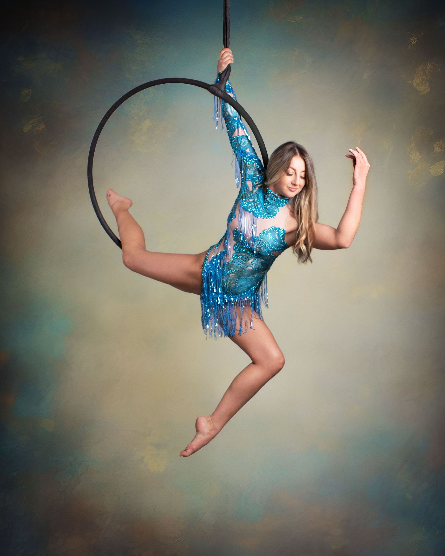 Aerial hoop performer in blue sequined leotard gracefully poses mid-air against a dreamy painted backdrop.