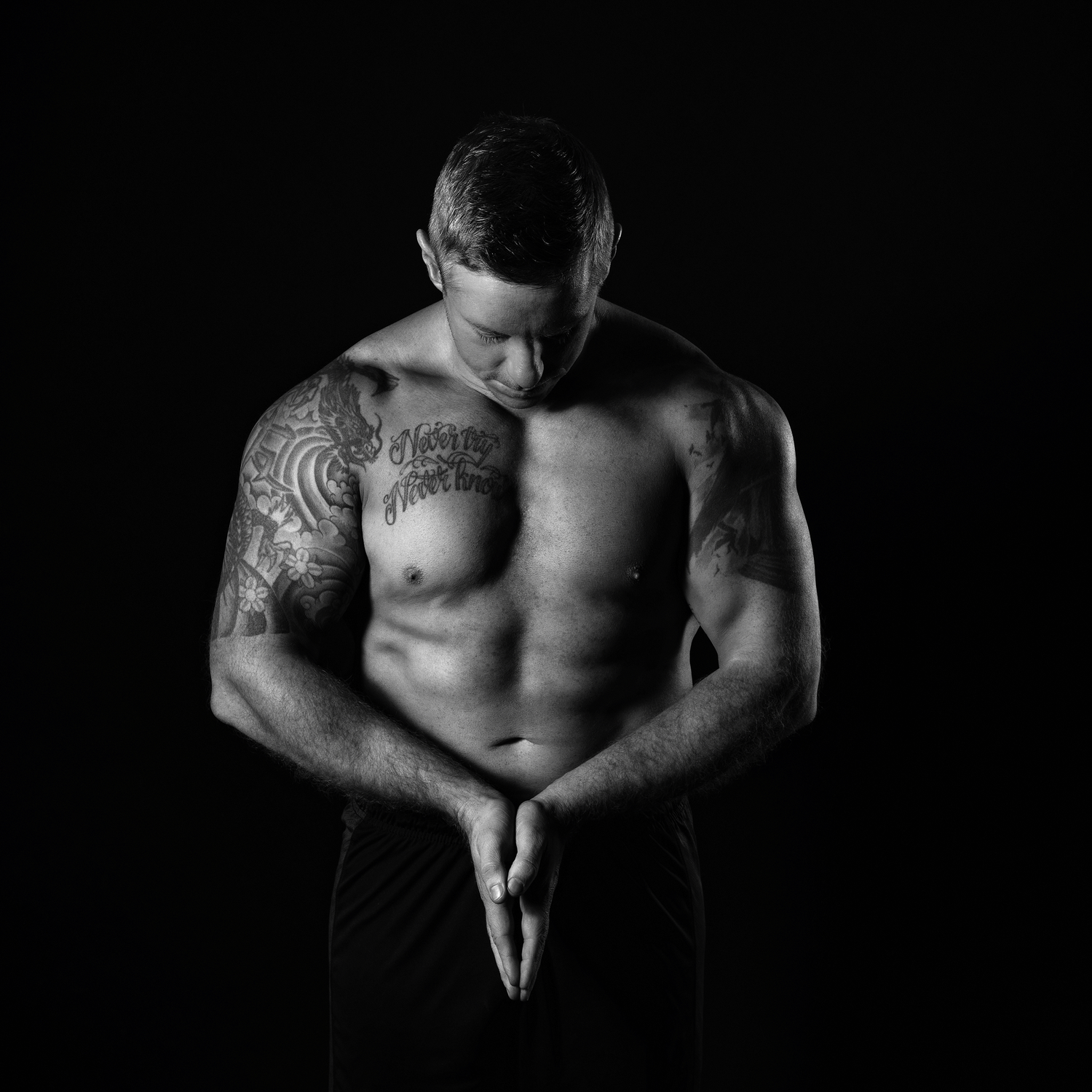 Black and white portrait of shirtless muscular person with tattoos standing in contemplative pose against dark background.