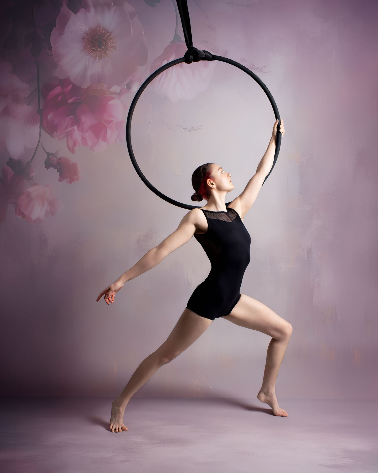 Aerial dancer in black leotard performs graceful poses on suspended hoop against dreamy pink floral backdrop.