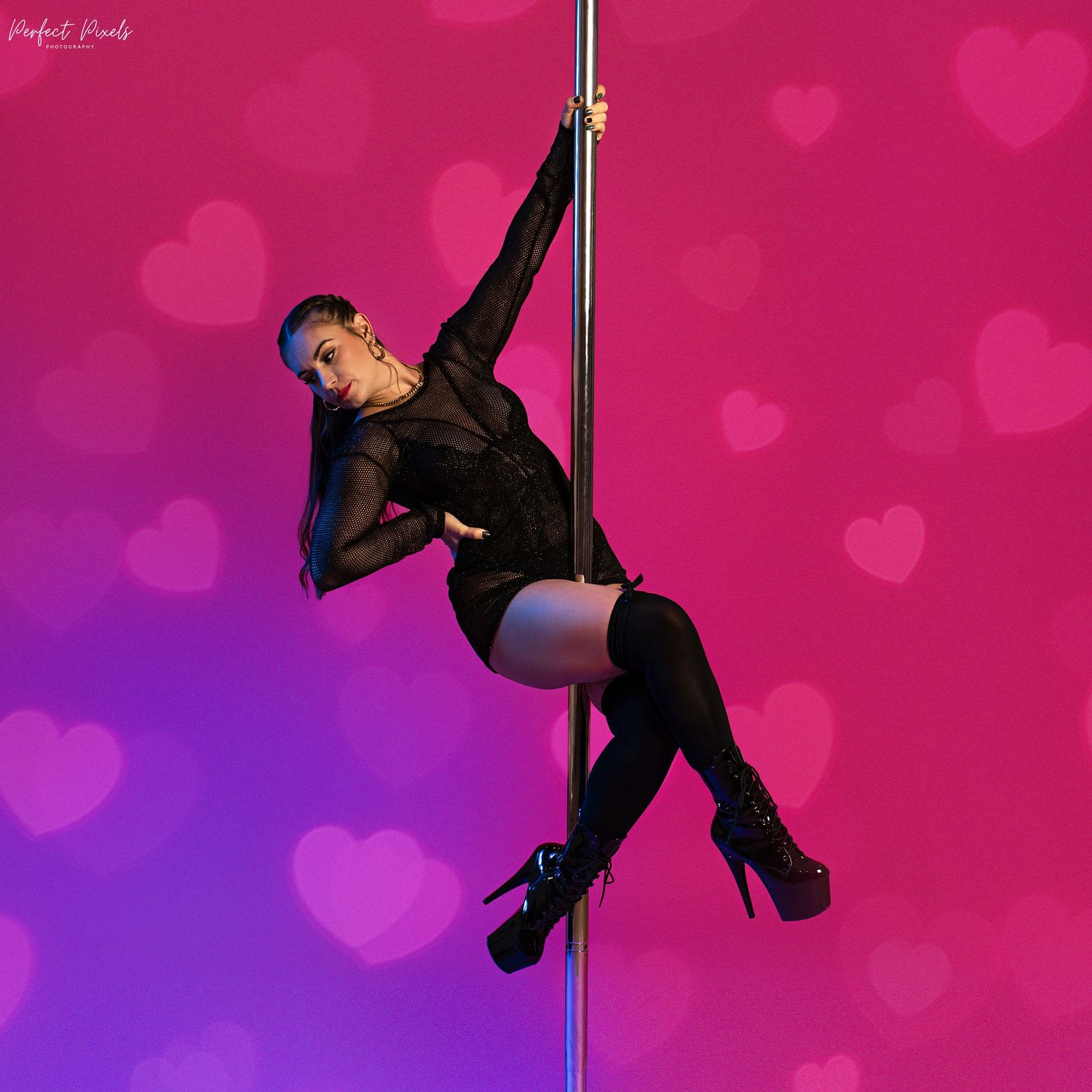 Athletic dancer performs pole routine in black outfit against pink heart backdrop.