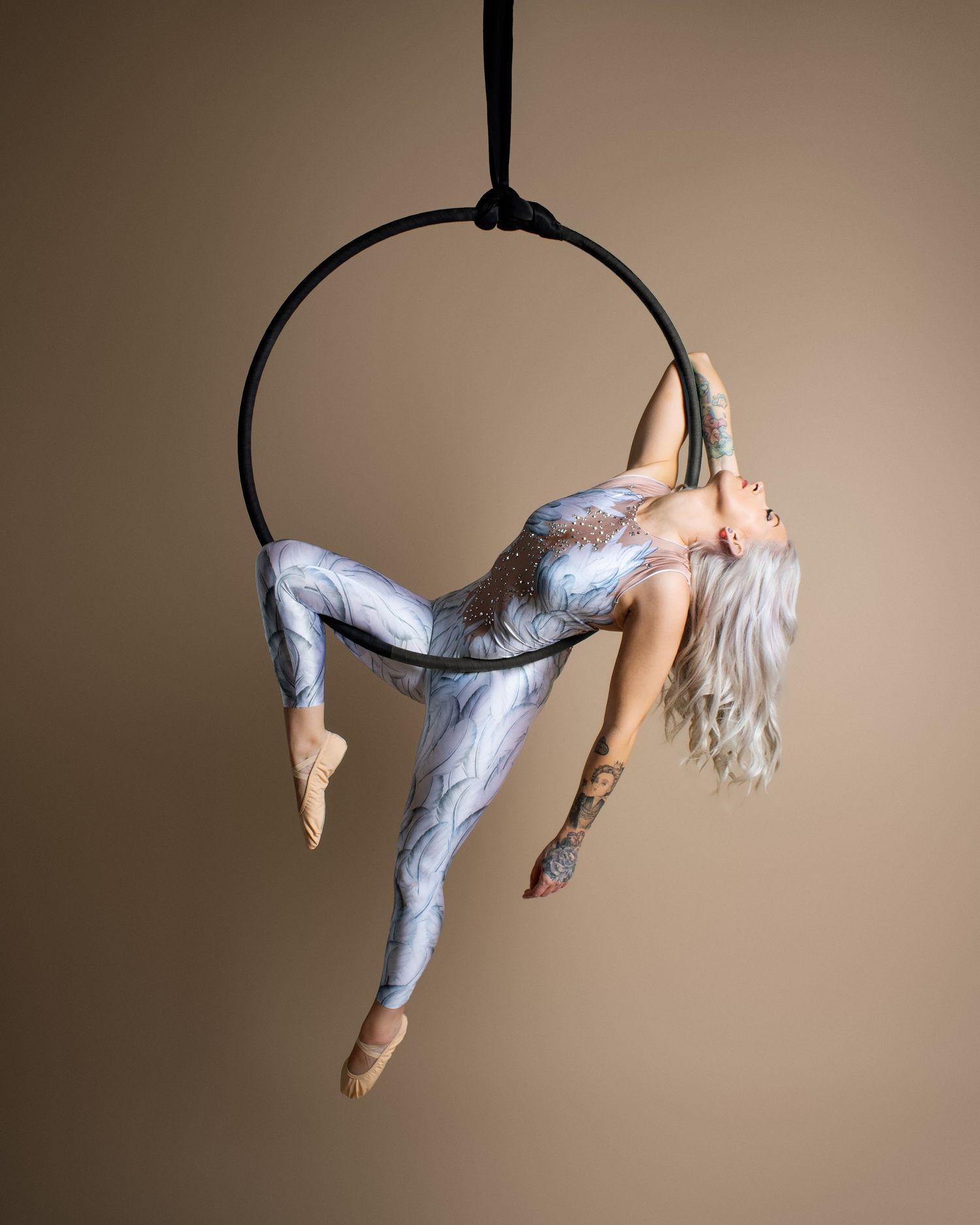 An aerial dancer performs a graceful sequence on a lyra hoop against a neutral brown backdrop.