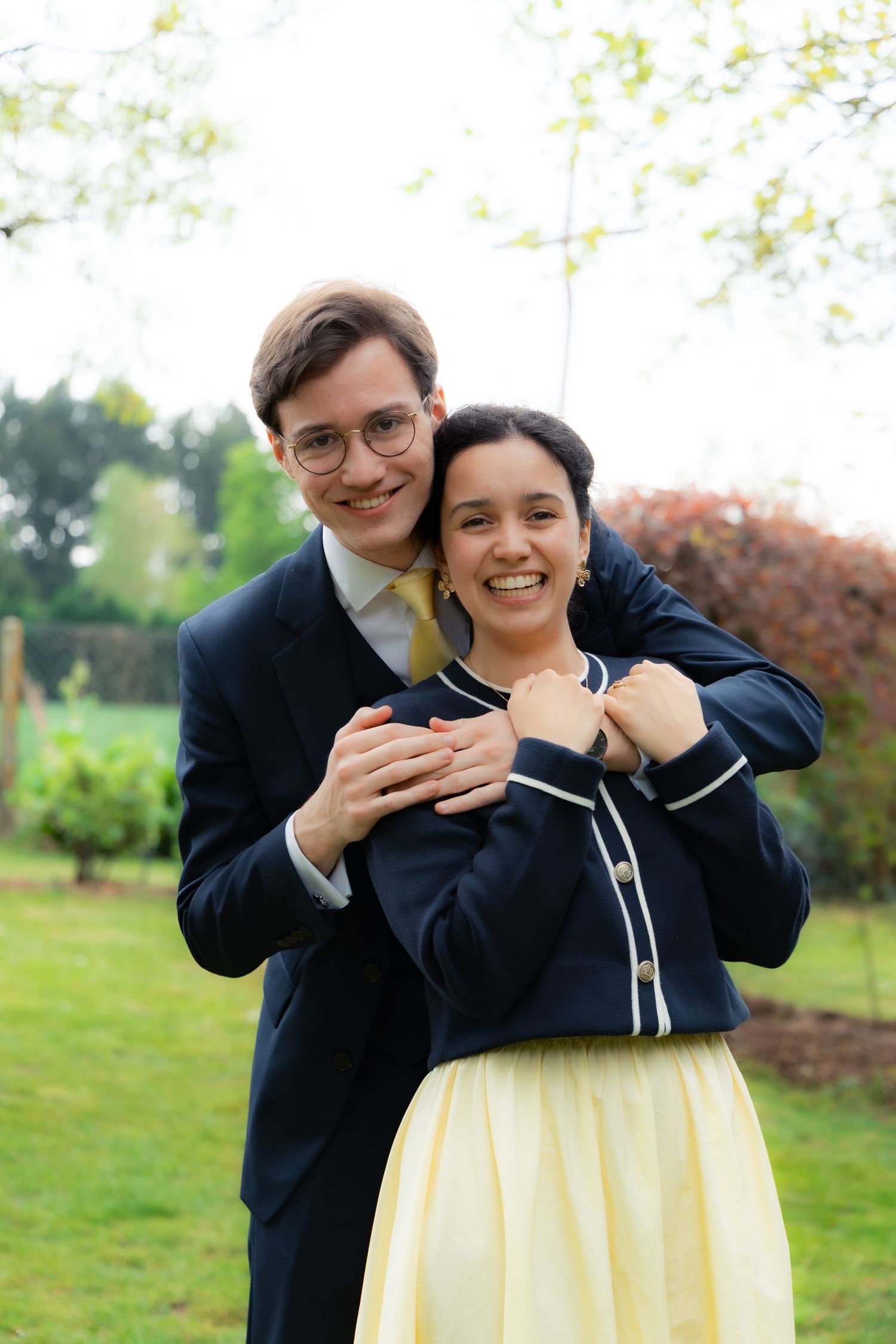 A smiling couple poses outdoors, man in navy suit hugging woman in yellow skirt and navy cardigan, in a lush garden.