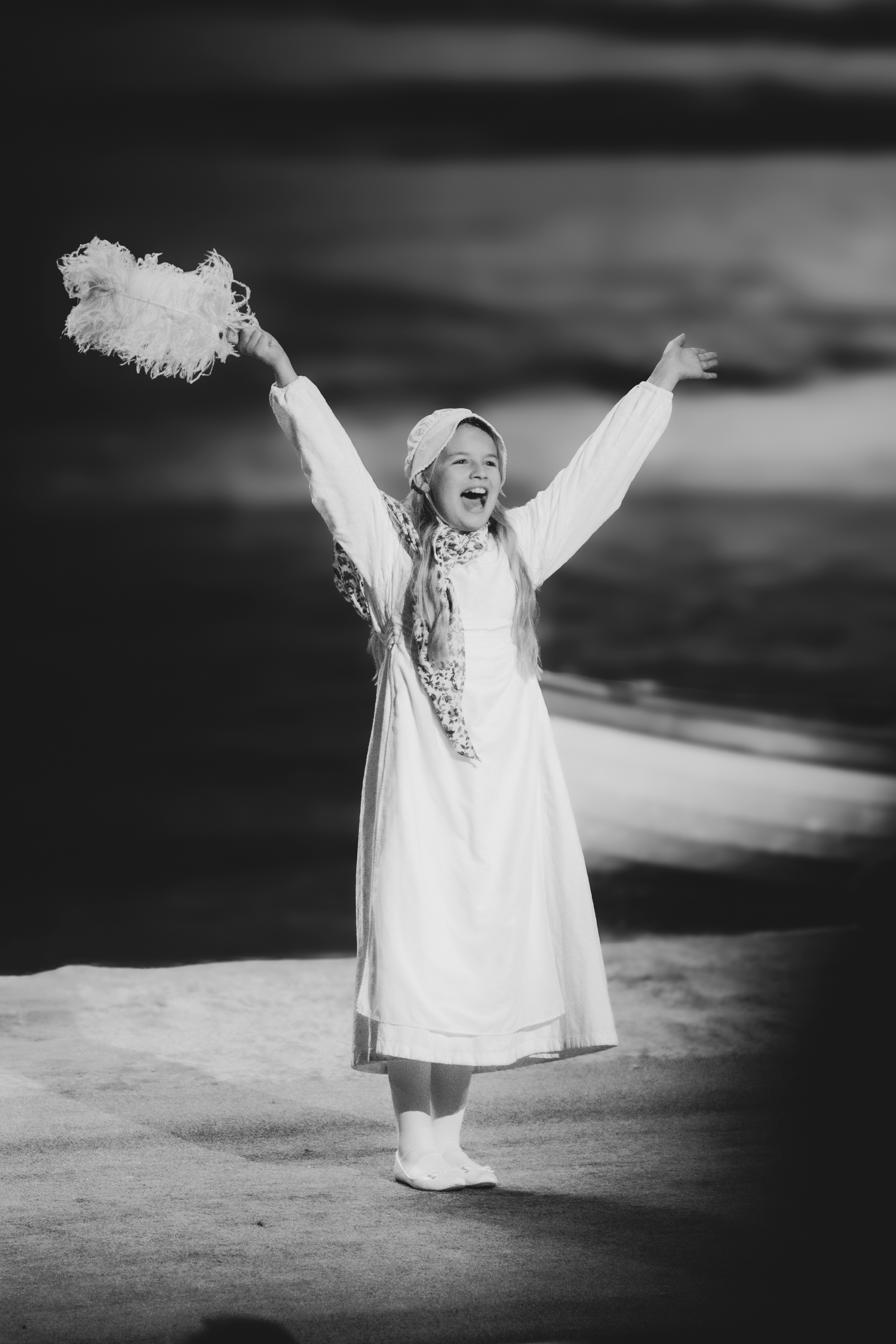 Black and white photo of a joyful girl in white dress and bonnet, arms raised, holding a feather fan on stage.