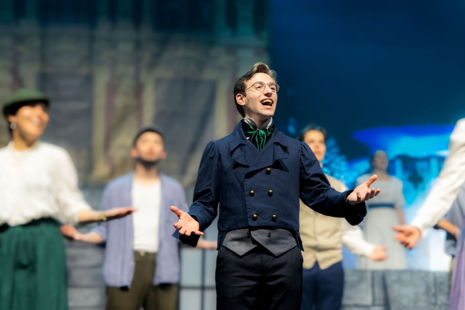 A young male performer sings on stage in a navy coat during a theatrical musical production.