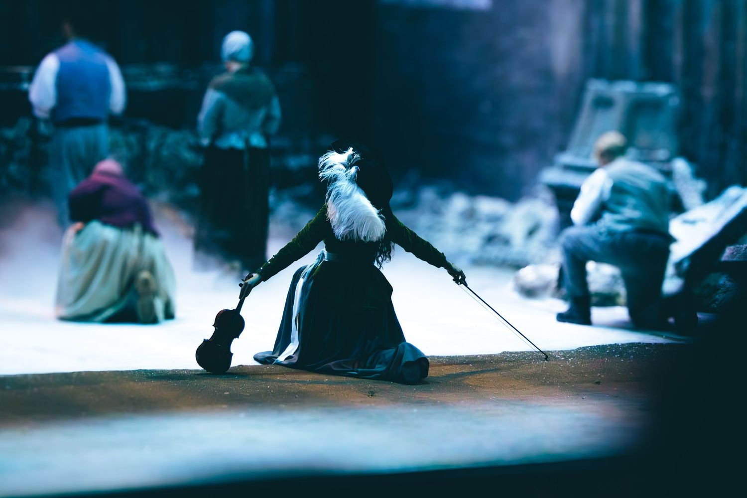 A dramatic theatrical performance featuring a costumed figure in a dark gown holding a violin bow on a misty stage.